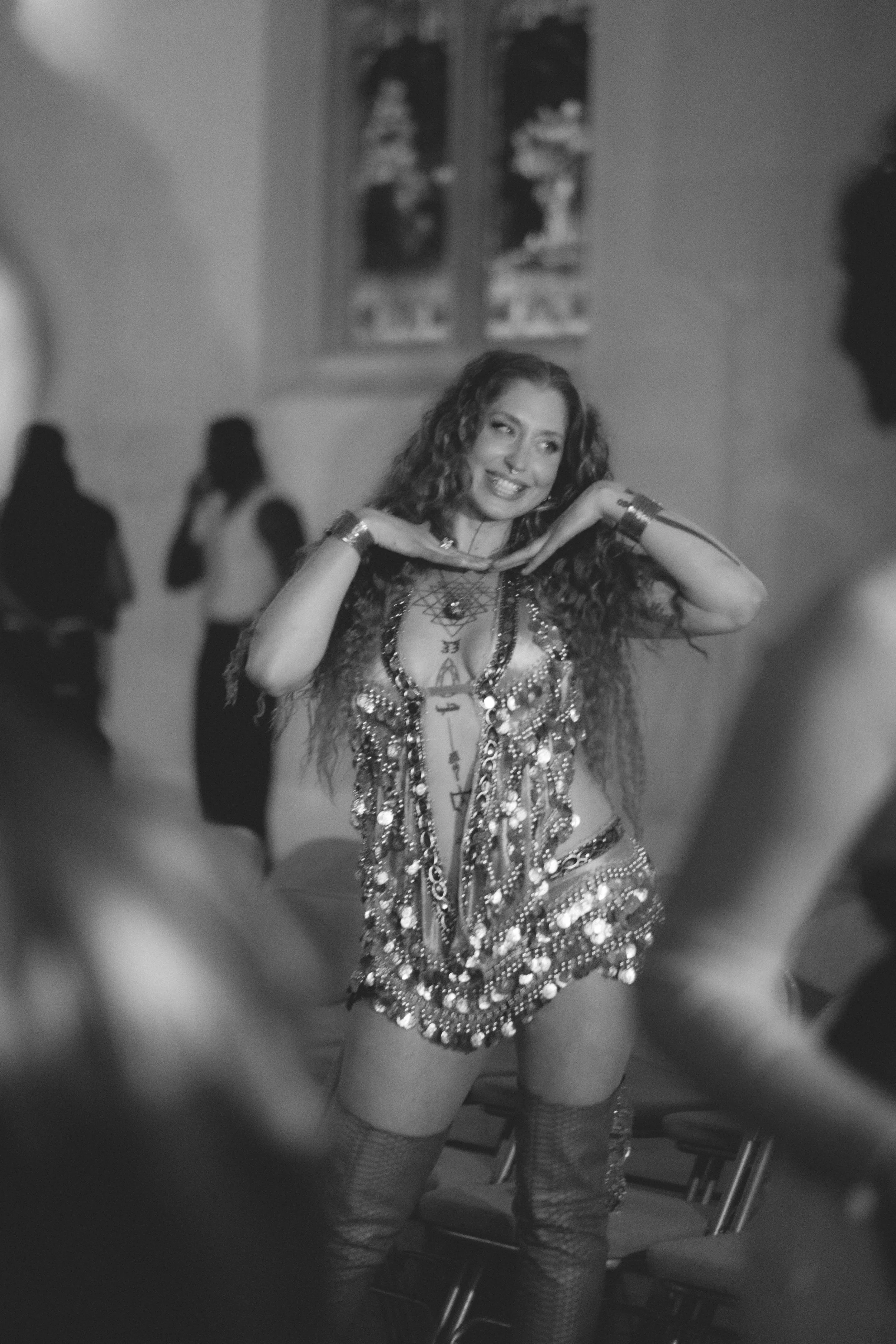 A woman in a sparkly, fringed belly dance costume posing with her hands under her chin, smiling, in an indoor setting with artwork on the wall and blurred figures in the background.