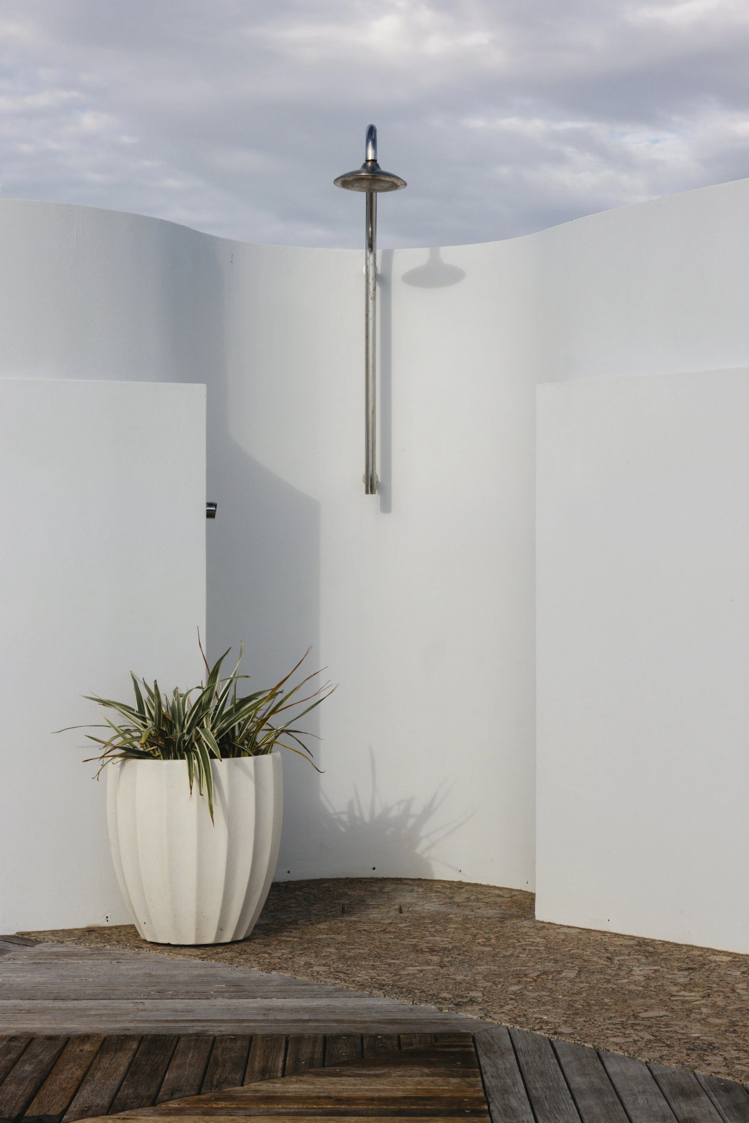 A white, curved wall with a metal outdoor shower head mounted on it, a white large pot with a green and white striped plant, and a wooden deck with a mat or rug. The scene is outdoors with a cloudy sky overhead.