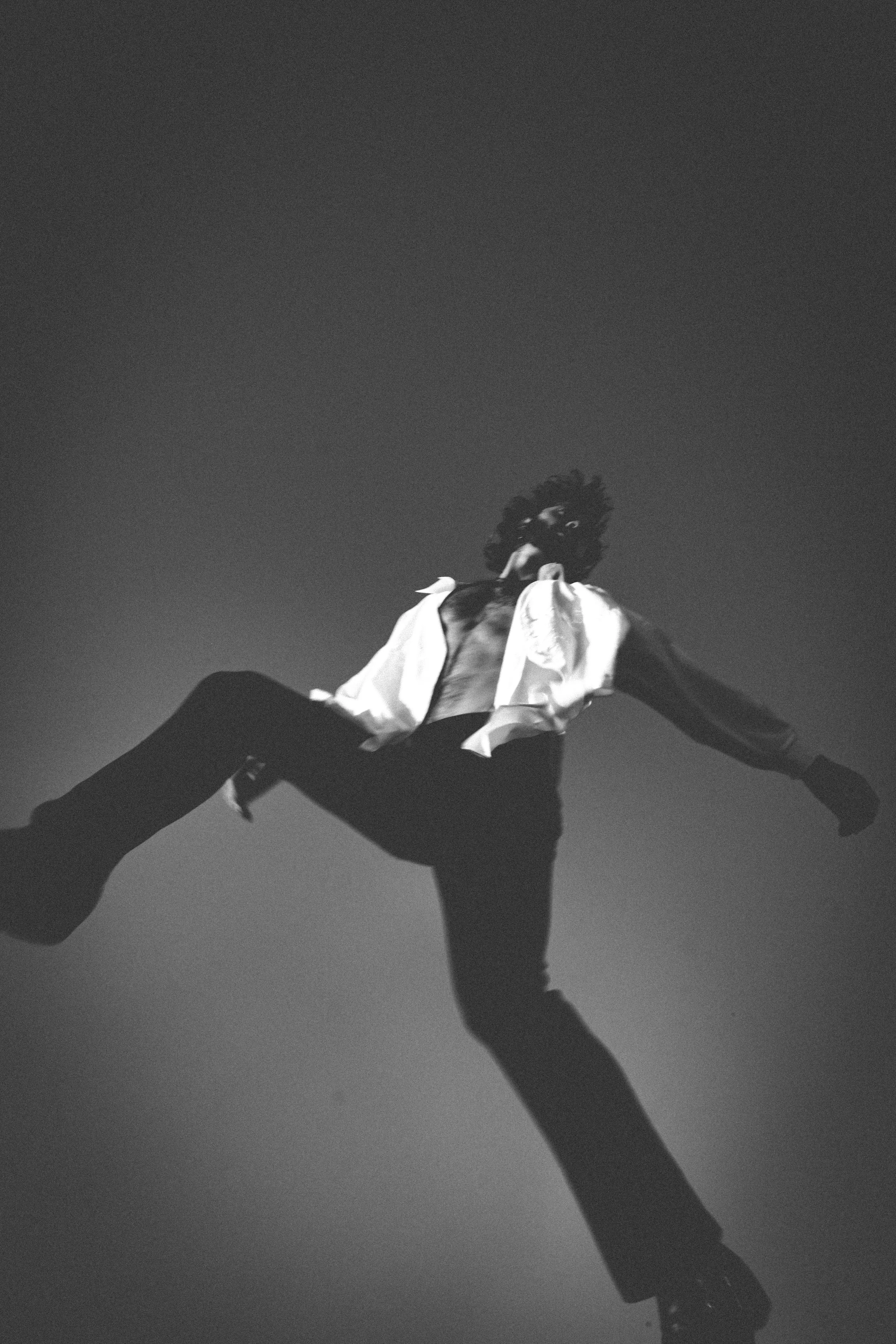 Black and white photo of a person in a white shirt and dark pants jumping or falling, with a clear sky background and their hair flying.