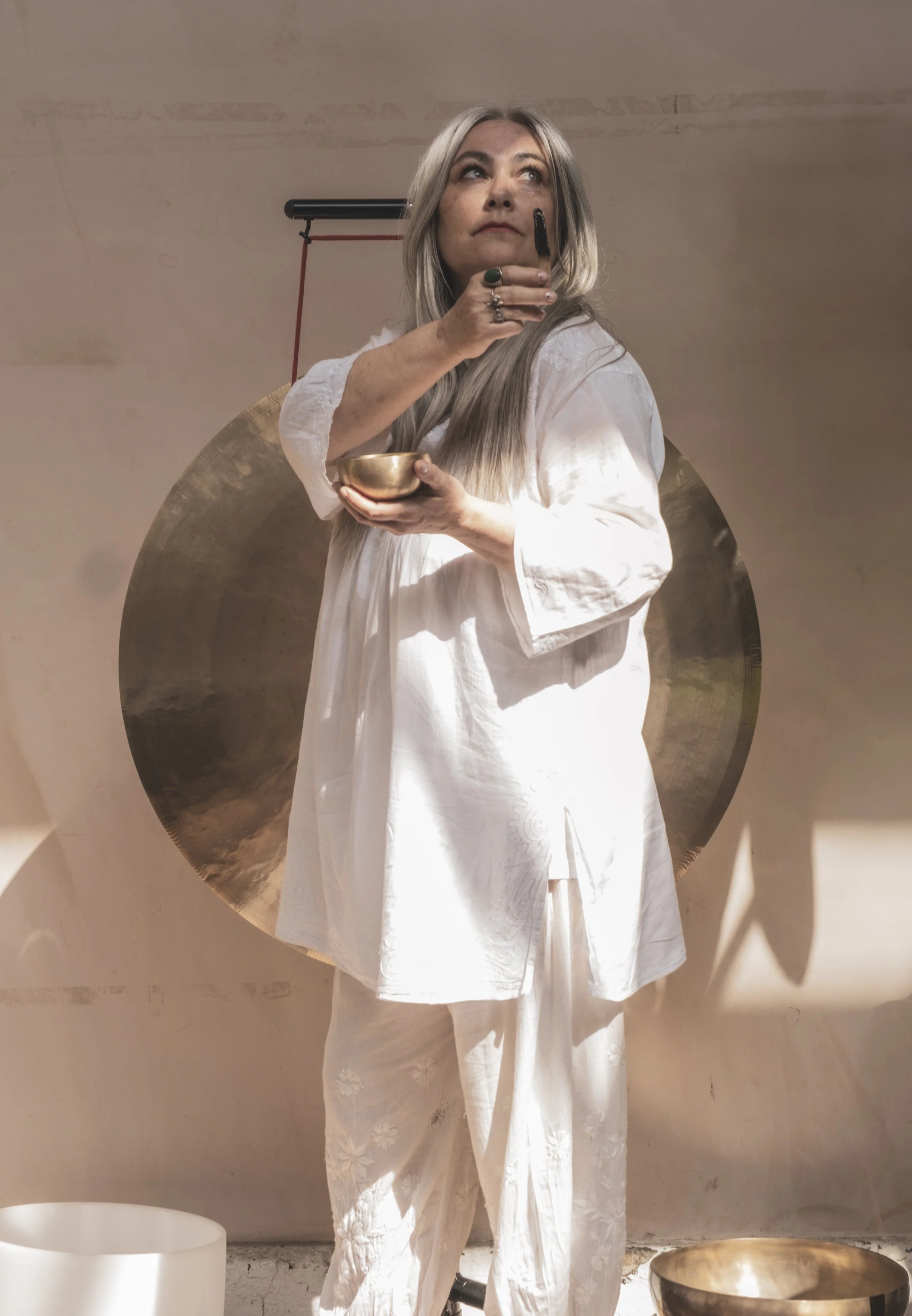 Woman with long, gray hair wearing a white dress holding a bell-shaped singing bowl and a small wooden mallet, standing in front of a large brass gong mounted on the wall.