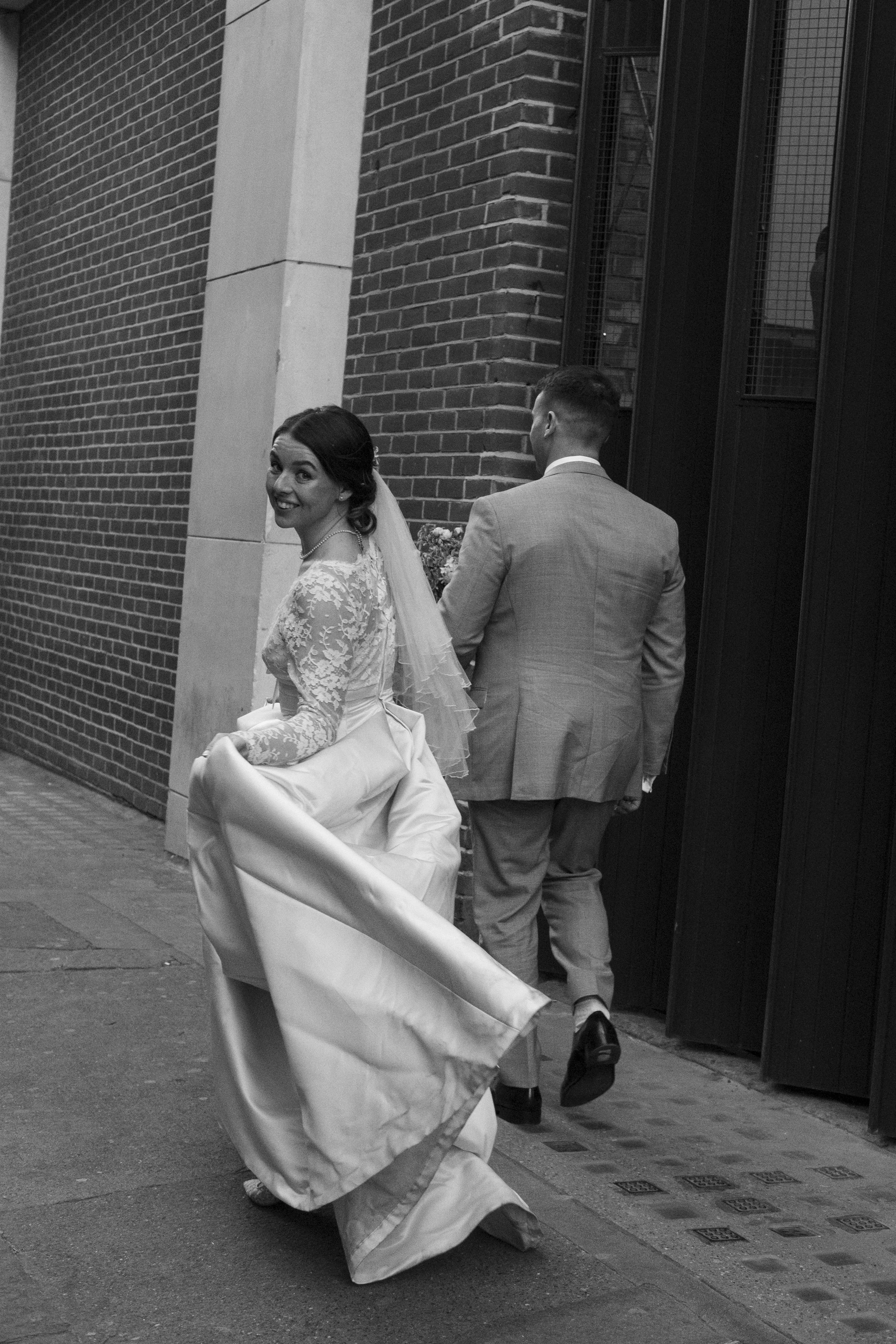 A bride in a lace wedding dress and veil is walking happily while holding up her dress with one hand, smiling at the camera. A groom wearing a light-colored suit is behind her, walking in the same direction near a brick wall and a metal gate.