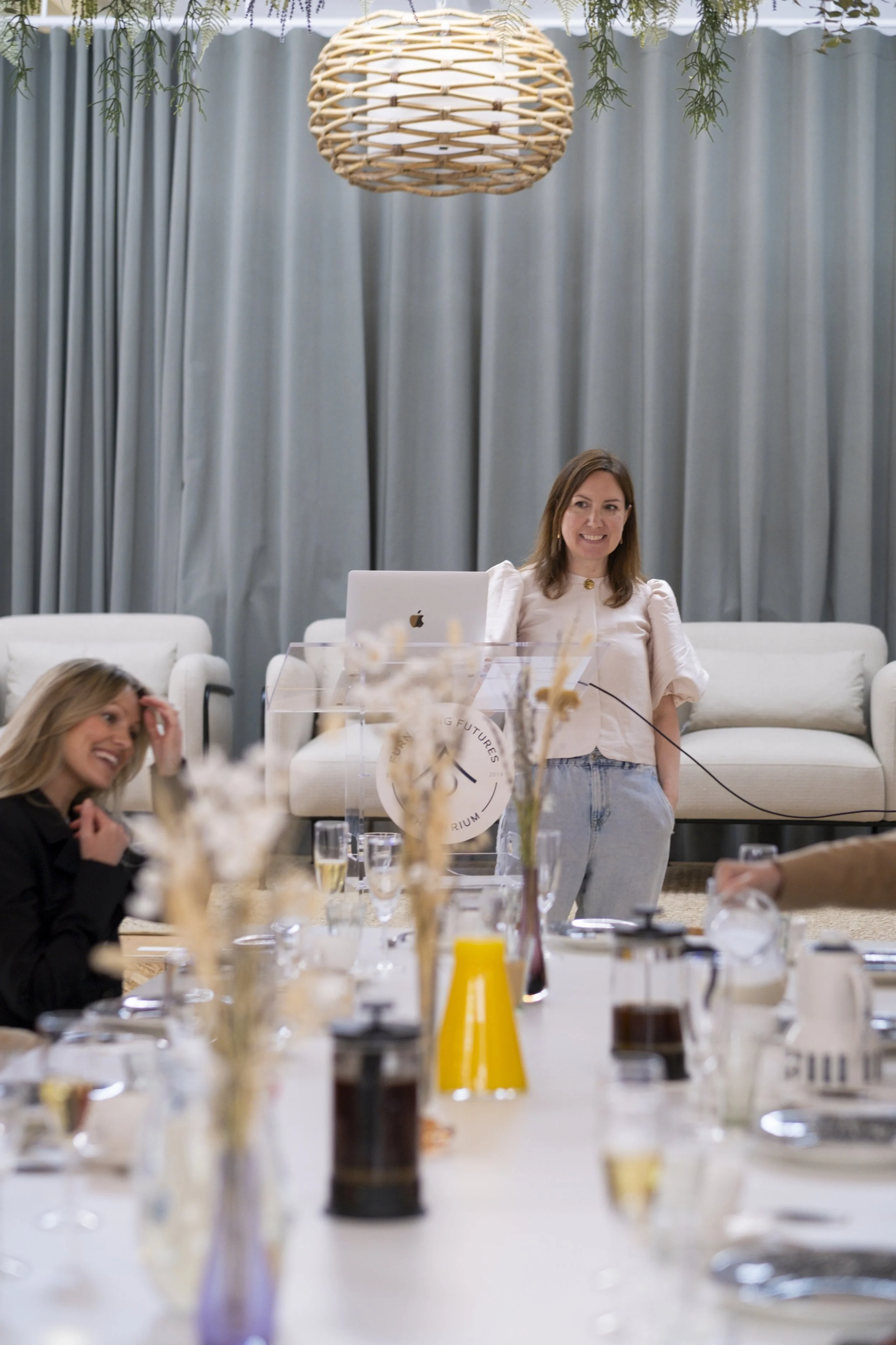 A woman standing behind a clear podium, smiling, with gray curtains and a wicker light fixture hanging from above. Seated people and a table with drinks and decorations are in the foreground.
