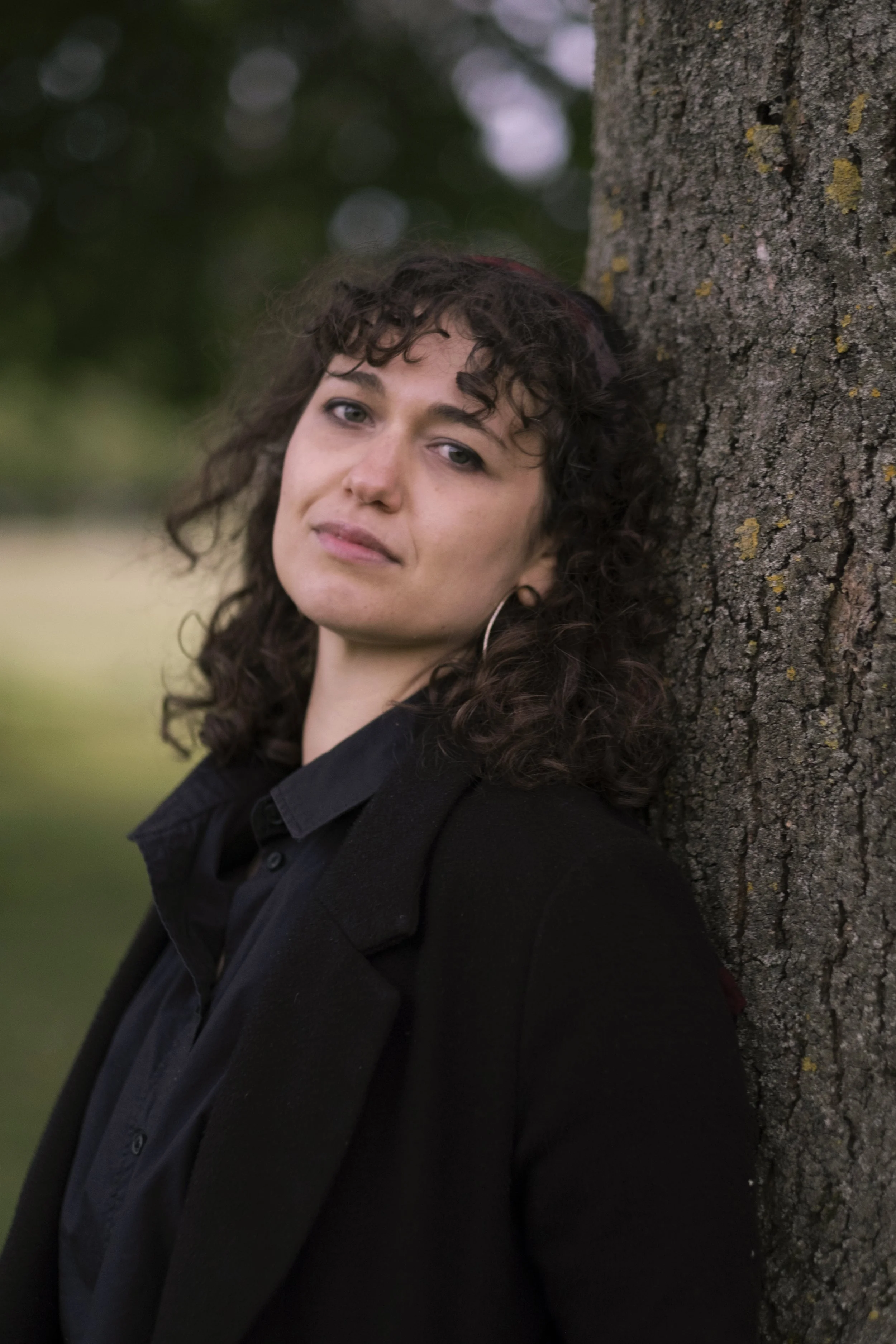 A woman with curly dark hair leaning against a tree outdoors, wearing a black jacket. Background is blurred with natural green and purple hues.