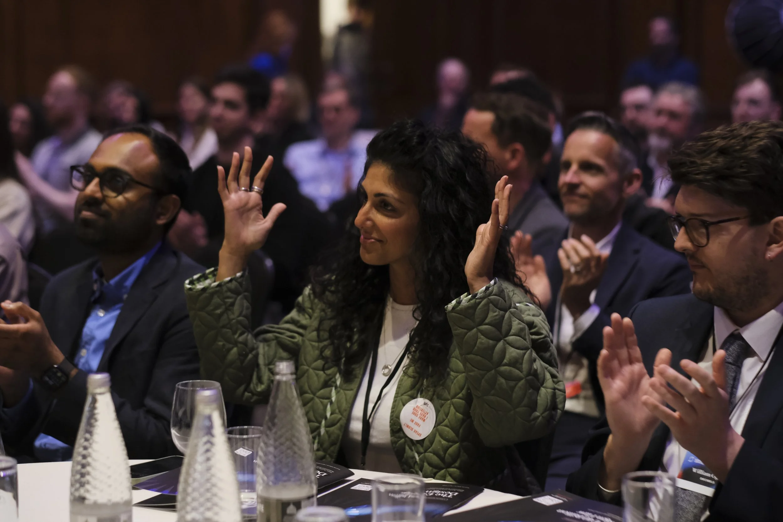 Audience attending a conference or seminar, with a diverse group of people seated at tables, some raising their hands, with glasses and bottles on the table.