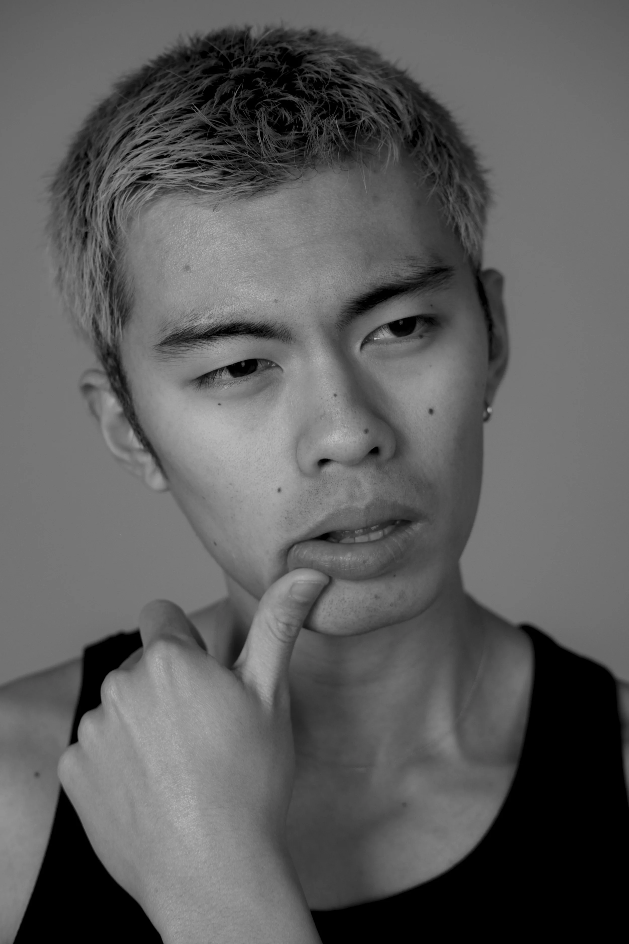 Close-up black-and-white photo of a young woman with short hair, touching her chin with her hand, wearing a sleeveless top.