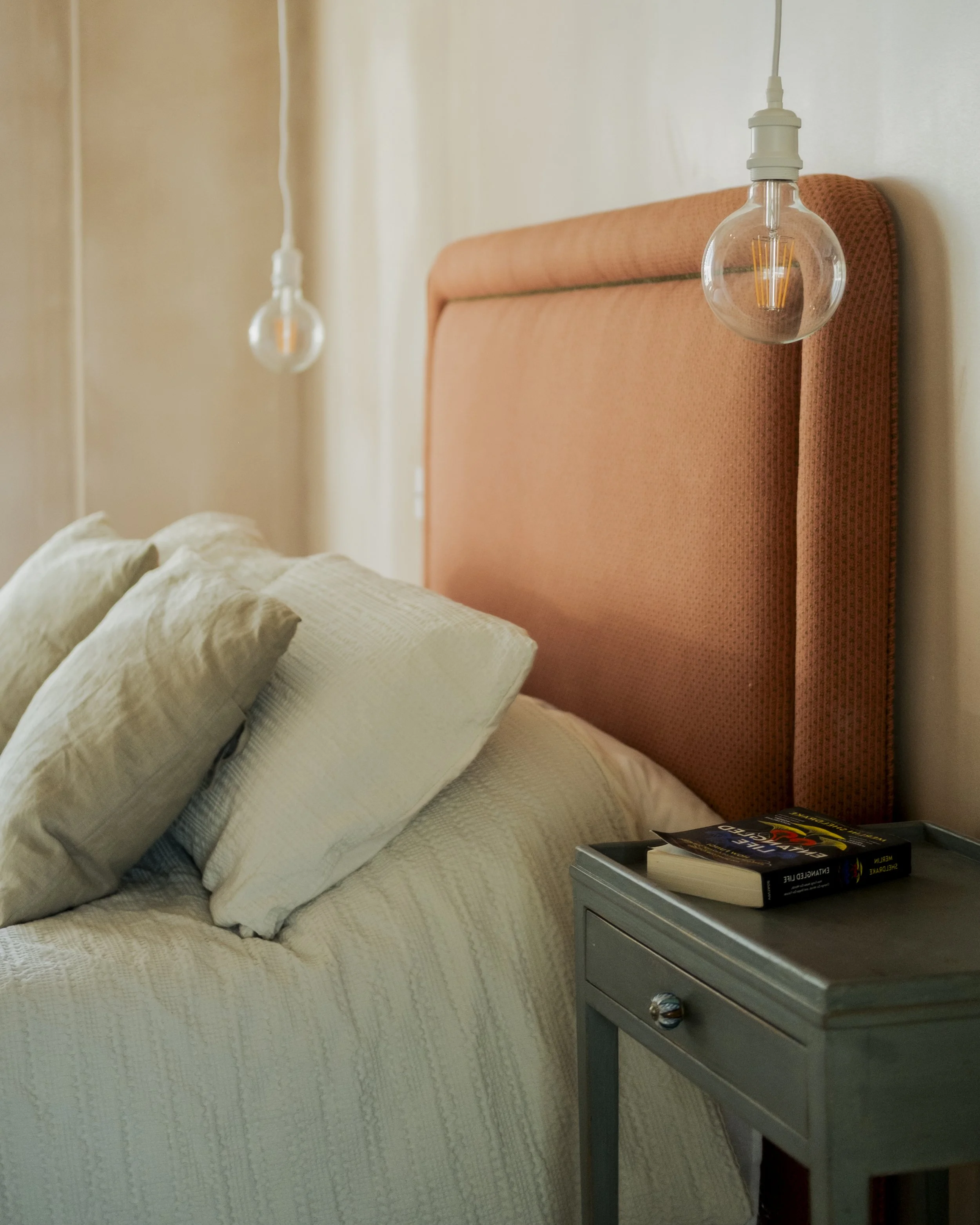 A cozy bedroom with a bed featuring beige bedding and multiple pillows, a mustard headboard, two glowing hanging light bulbs, and a gray nightstand with books on top.