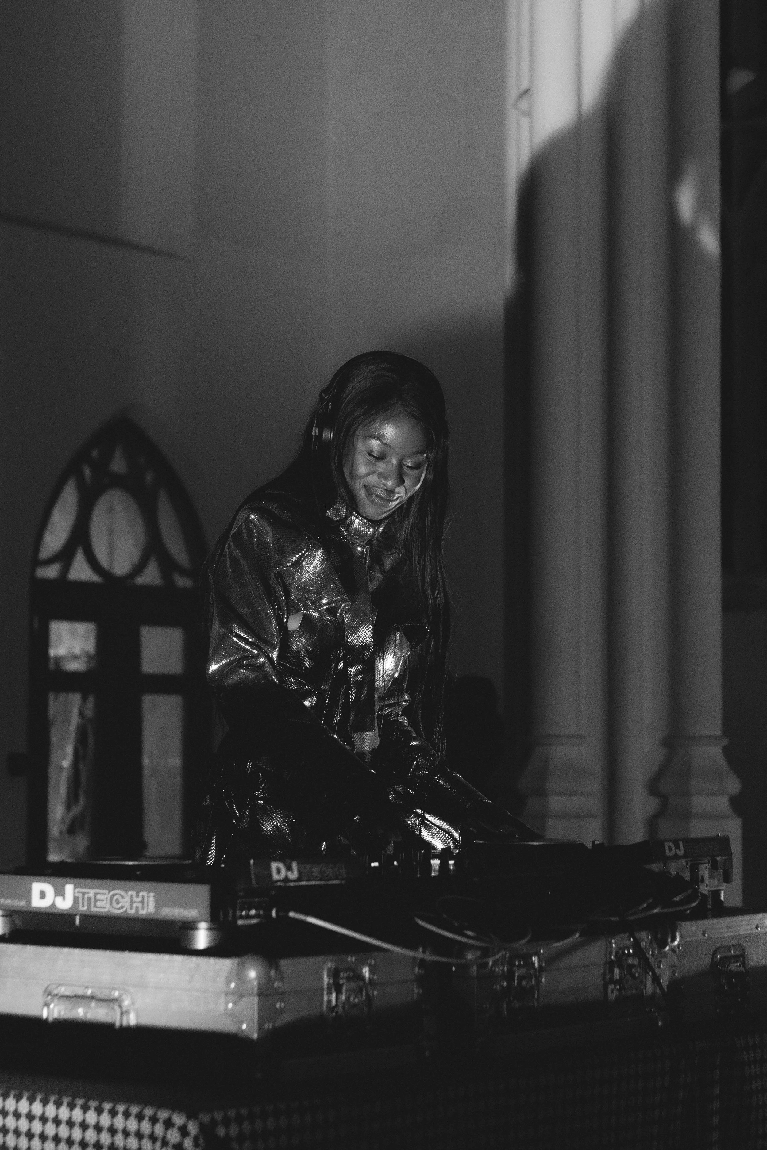 Black and white photo of a woman with long hair wearing headphones, smiling while DJing with turntables and mixer.