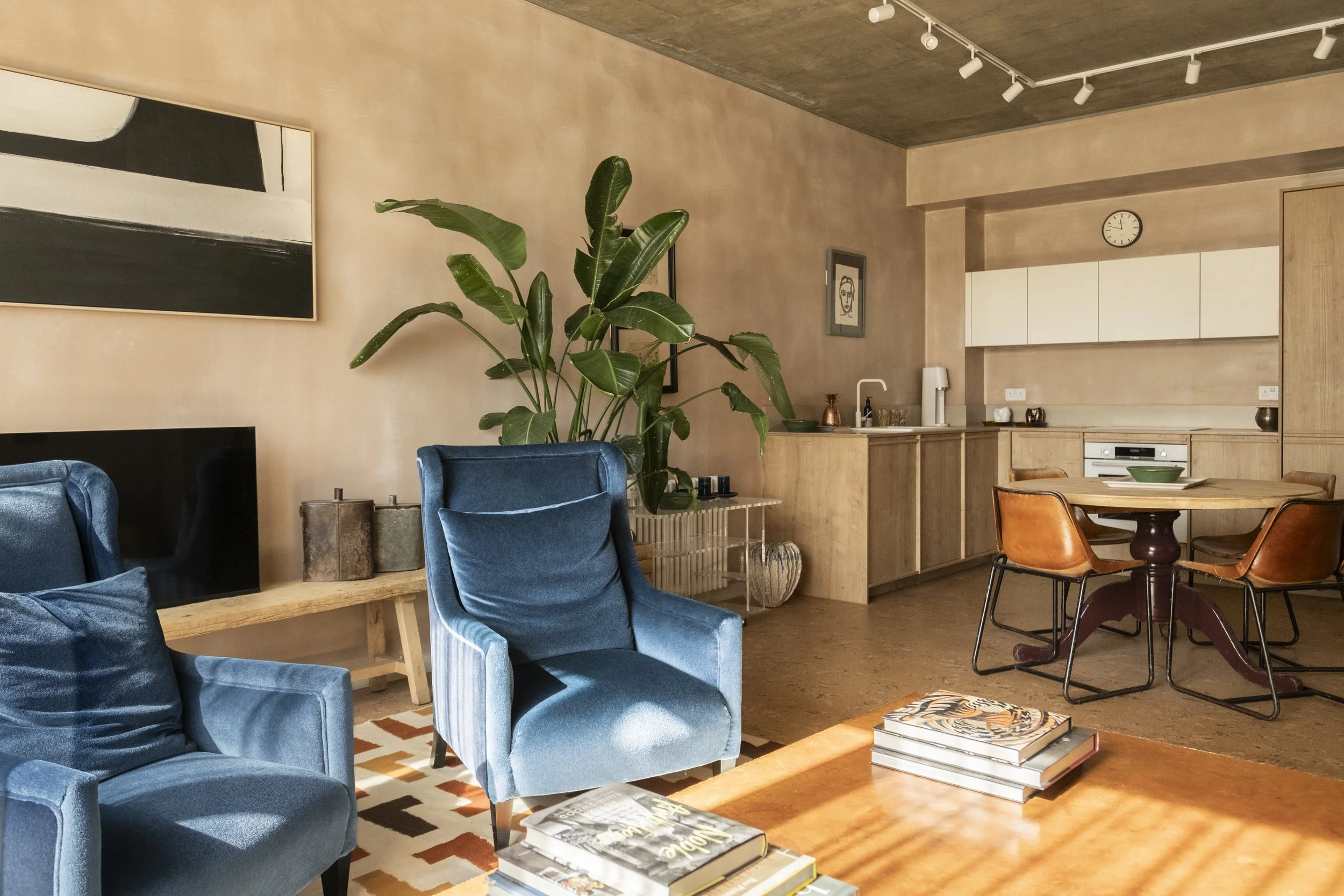 A modern living room with blue velvet armchairs, a large plant, artwork on the walls, and a kitchen area with white cabinets and a clock on the wall.