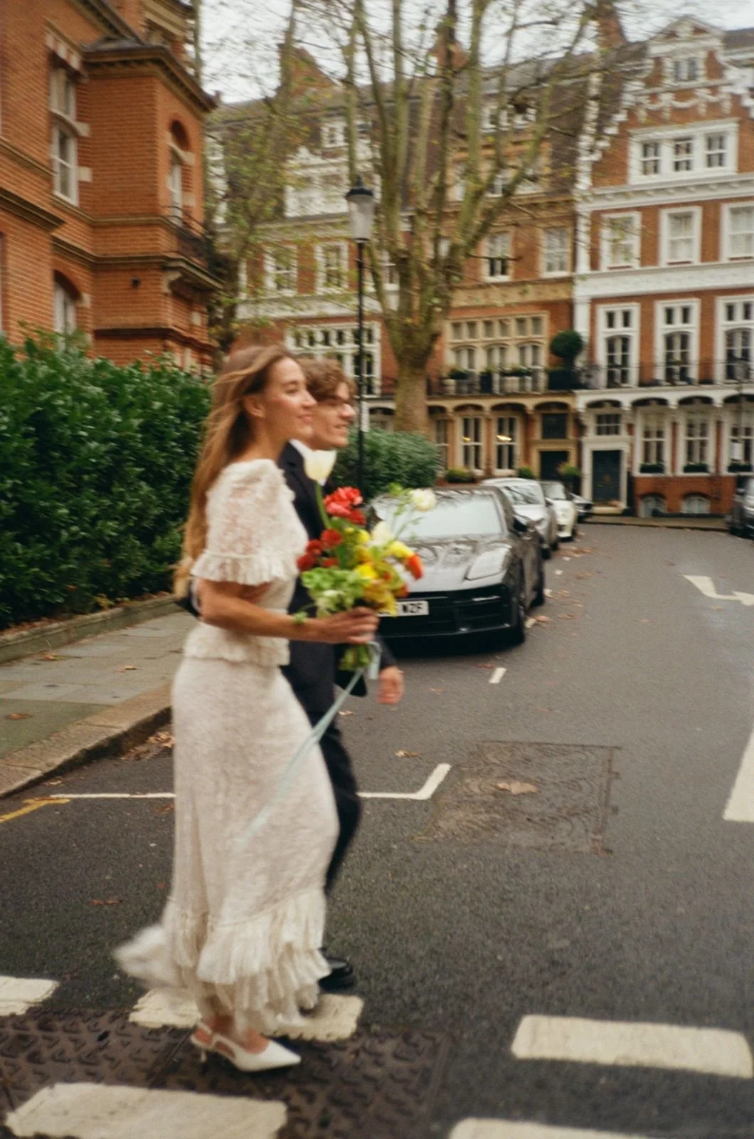 A woman in a white dress holding a bouquet of flowers walking on a city street, accompanied by a man in a tuxedo. They appear to be celebrating or attending a special event. The background features brick buildings, parked cars, and a tree.