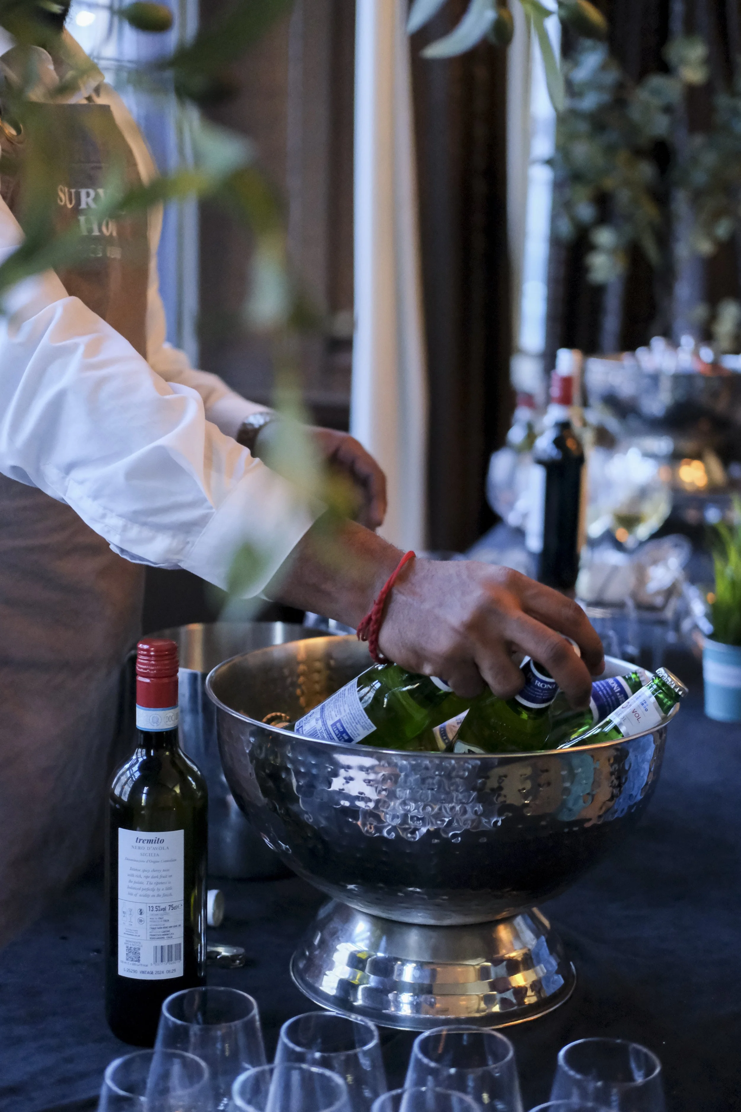 Person's hand reaching into a silver bowl filled with green glass bottles, with wine bottles and glasses on a dark table in the background.