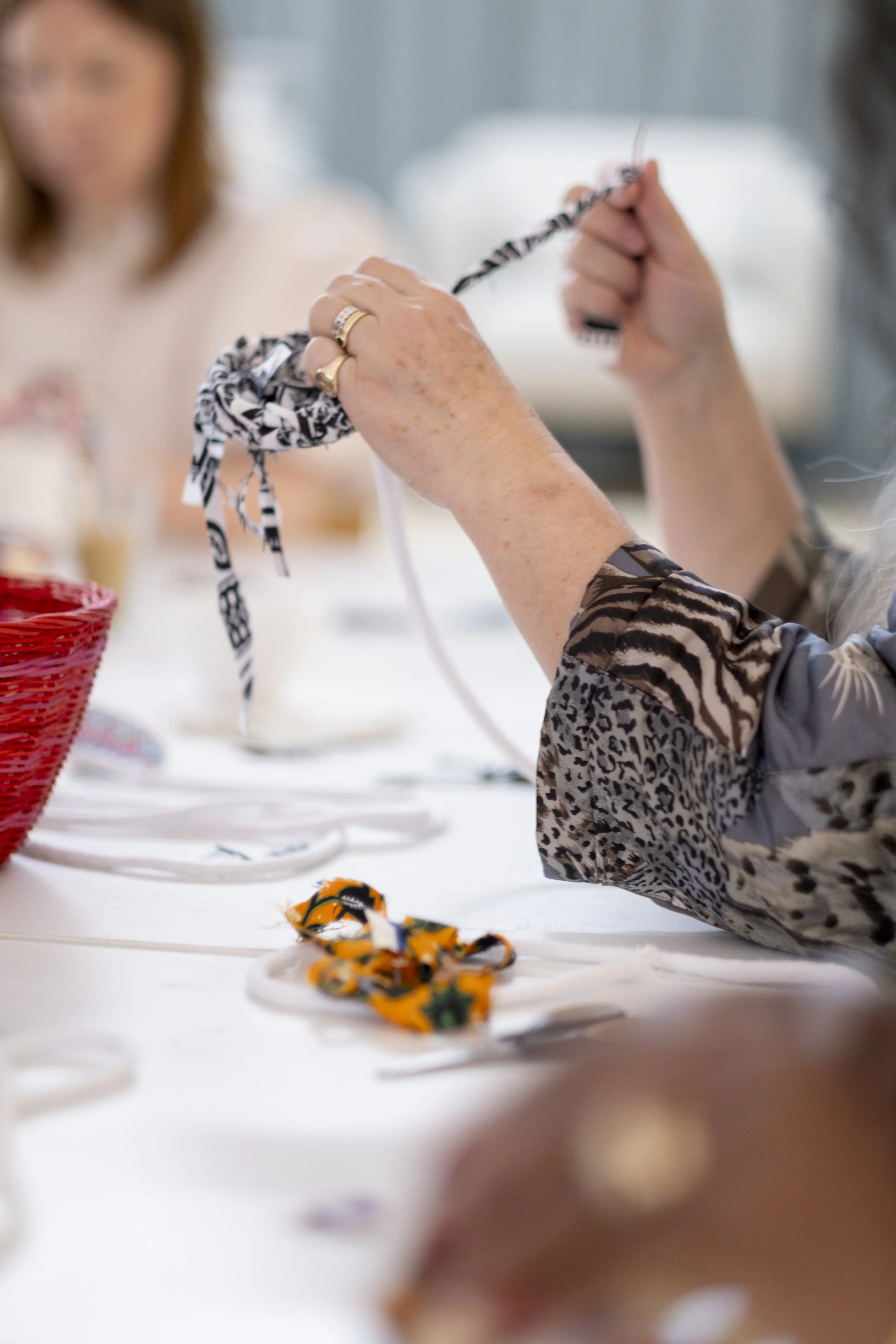 Close-up of a person's hands holding a fabric scrunchie with a black and white pattern. The person is wearing multiple rings. In the background, there is a blurred woman and some colorful fabric items on a white table.