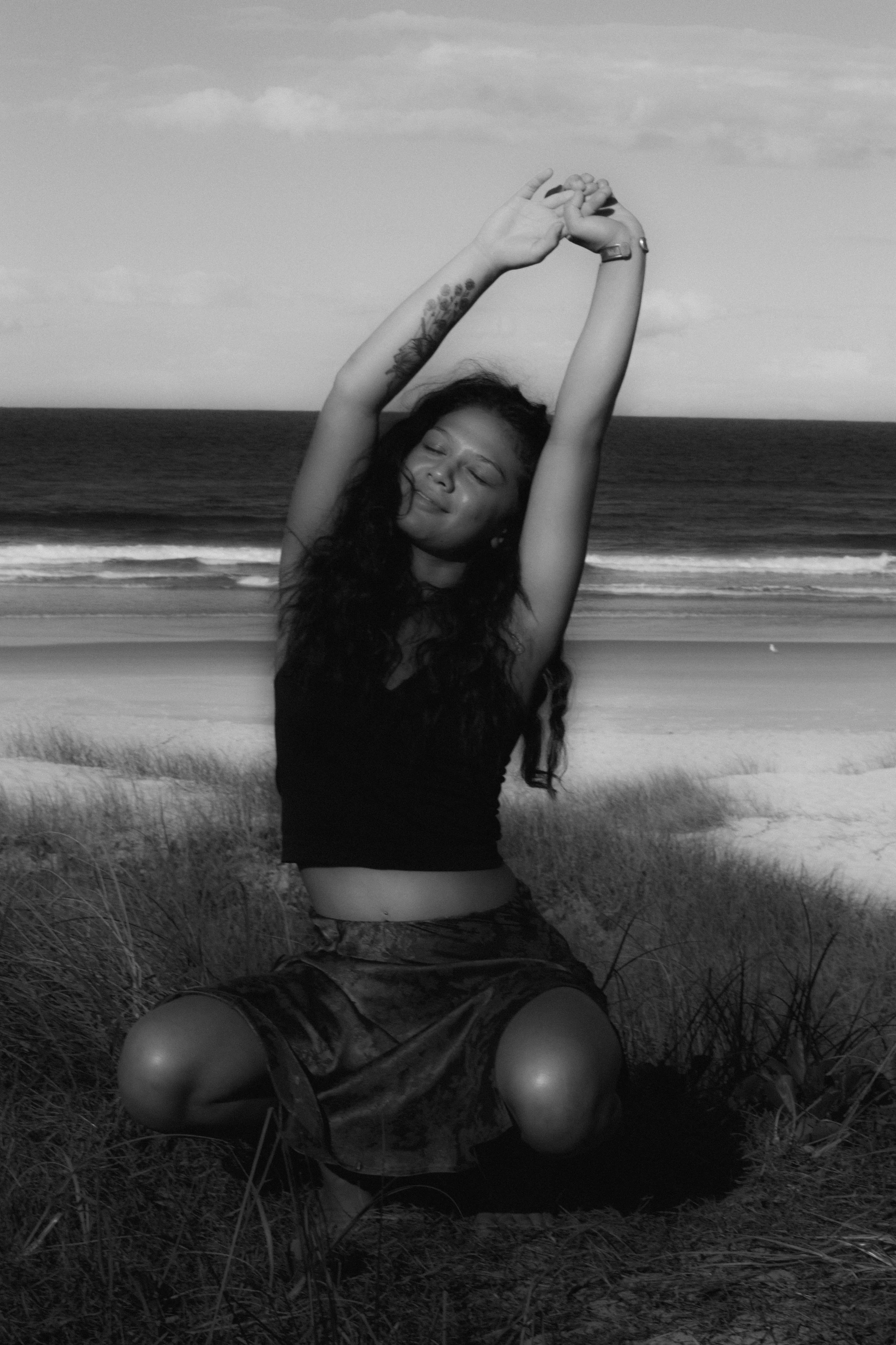 A woman with curly hair and tattoos on her arm is sitting outdoors on a grassy area near the beach. She has her arms raised above her head with her eyes closed and a peaceful expression on her face. The ocean and sky are visible in the background.