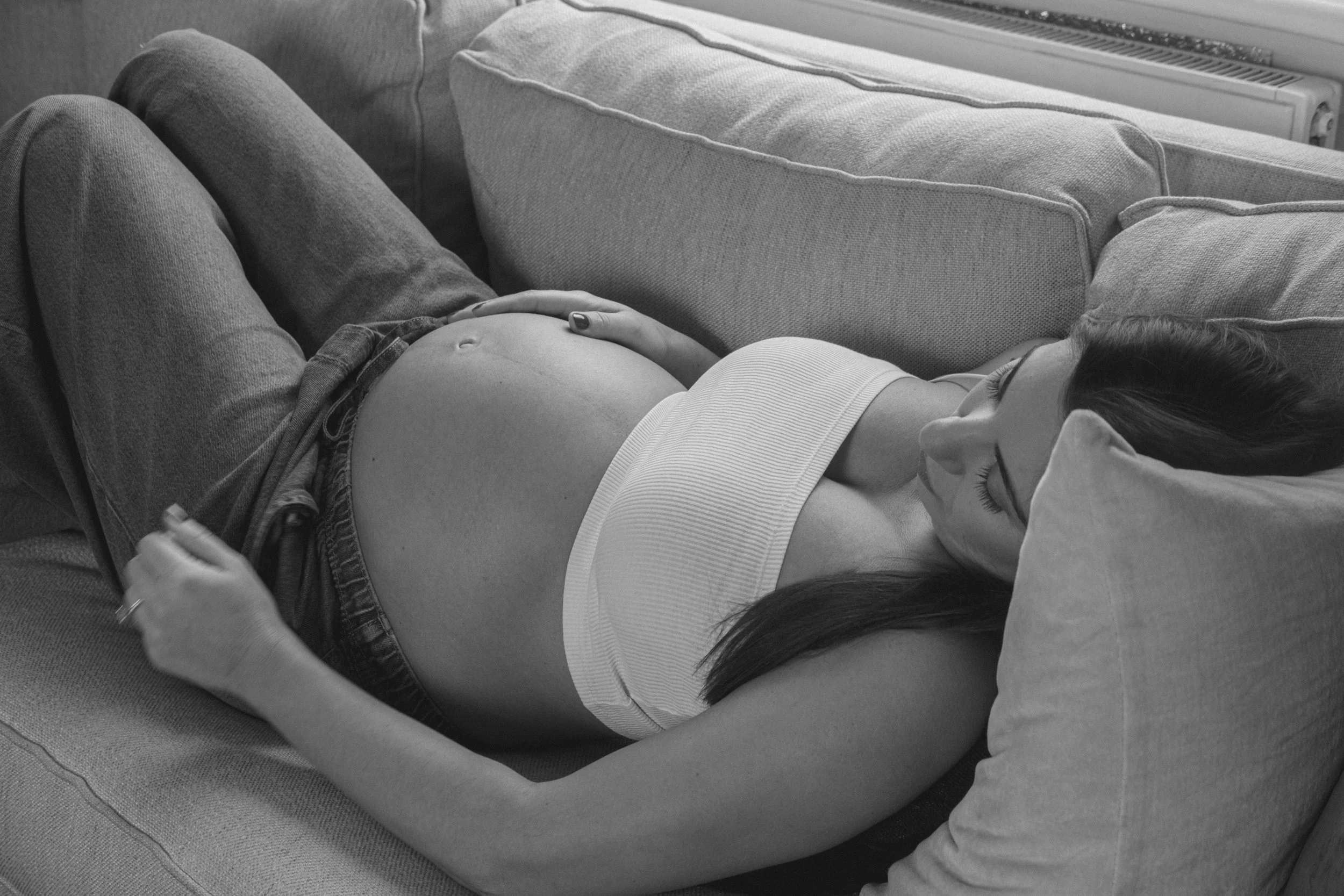 A woman lying on a couch with her hand on her pregnant belly, while a person sitting beside her holds her hand and drinks from a glass.