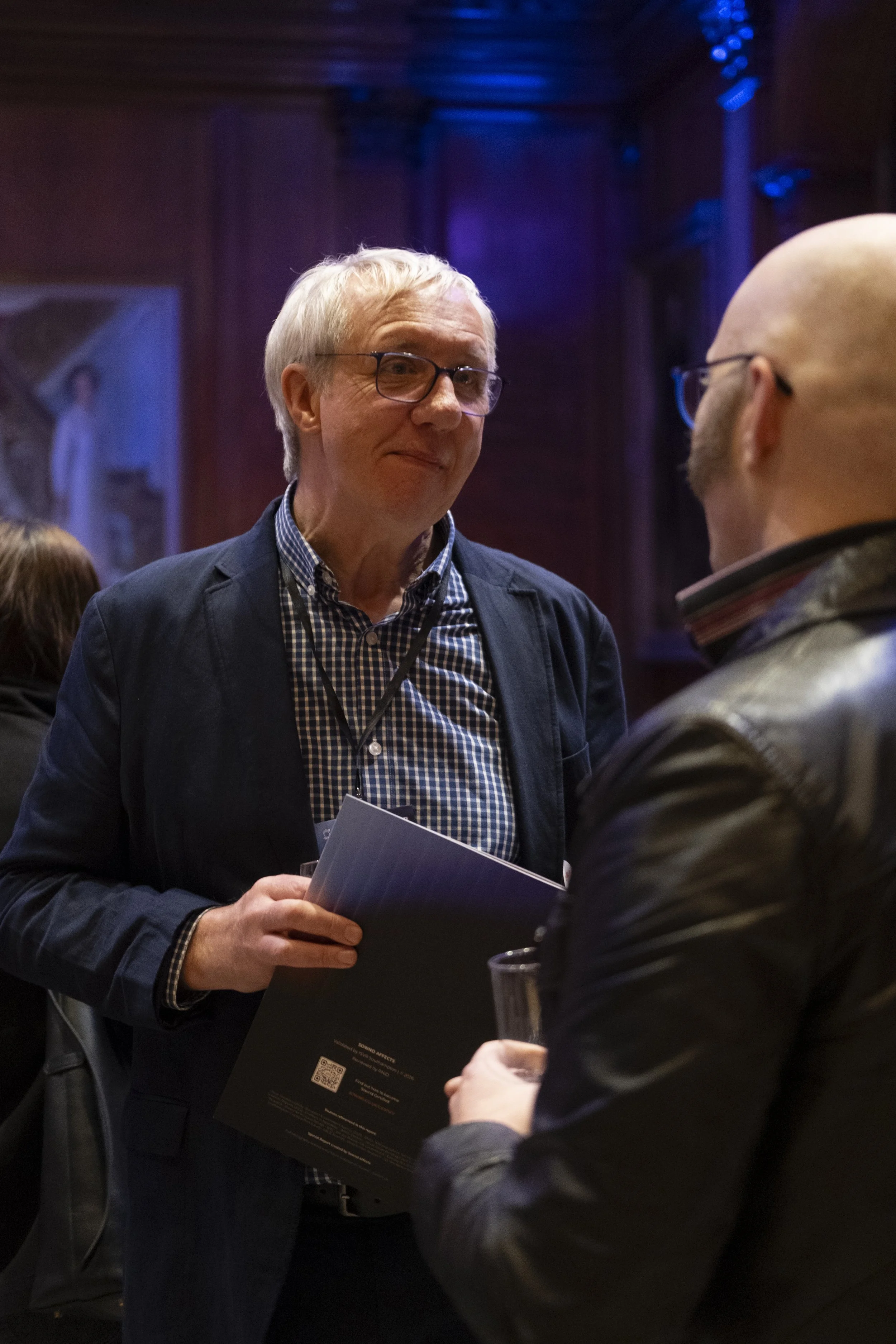 Two men are engaged in a conversation at a social event. One man with gray hair and glasses is holding a booklet, while the other, who is bald, is holding a glass. The setting appears to be indoors with dim lighting.