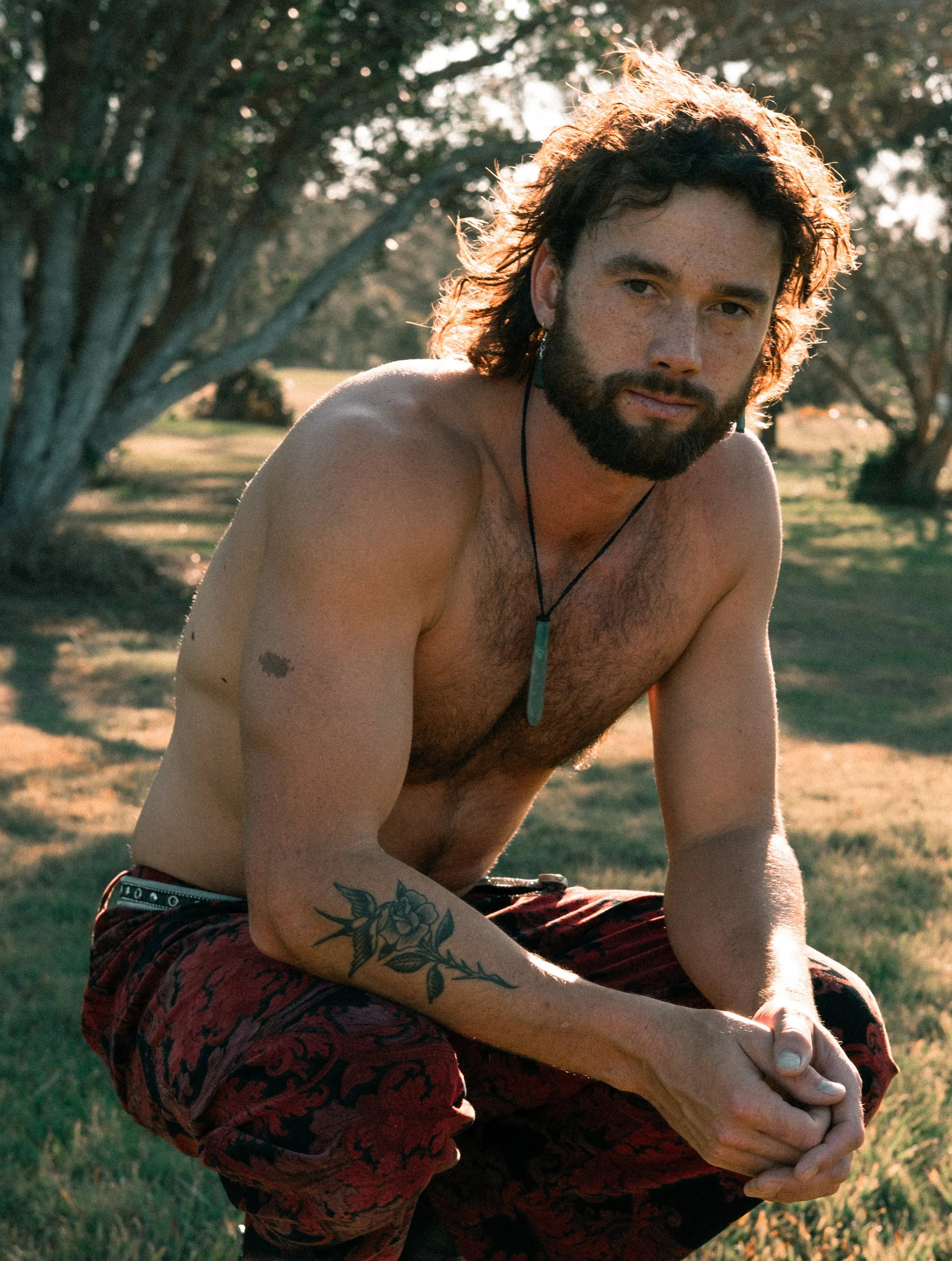 A shirtless man with curly hair and a beard, sitting outdoors during golden hour, wearing patterned red shorts and a turquoise pendant necklace, with tattoos on his arm, in a grassy area with trees in the background.