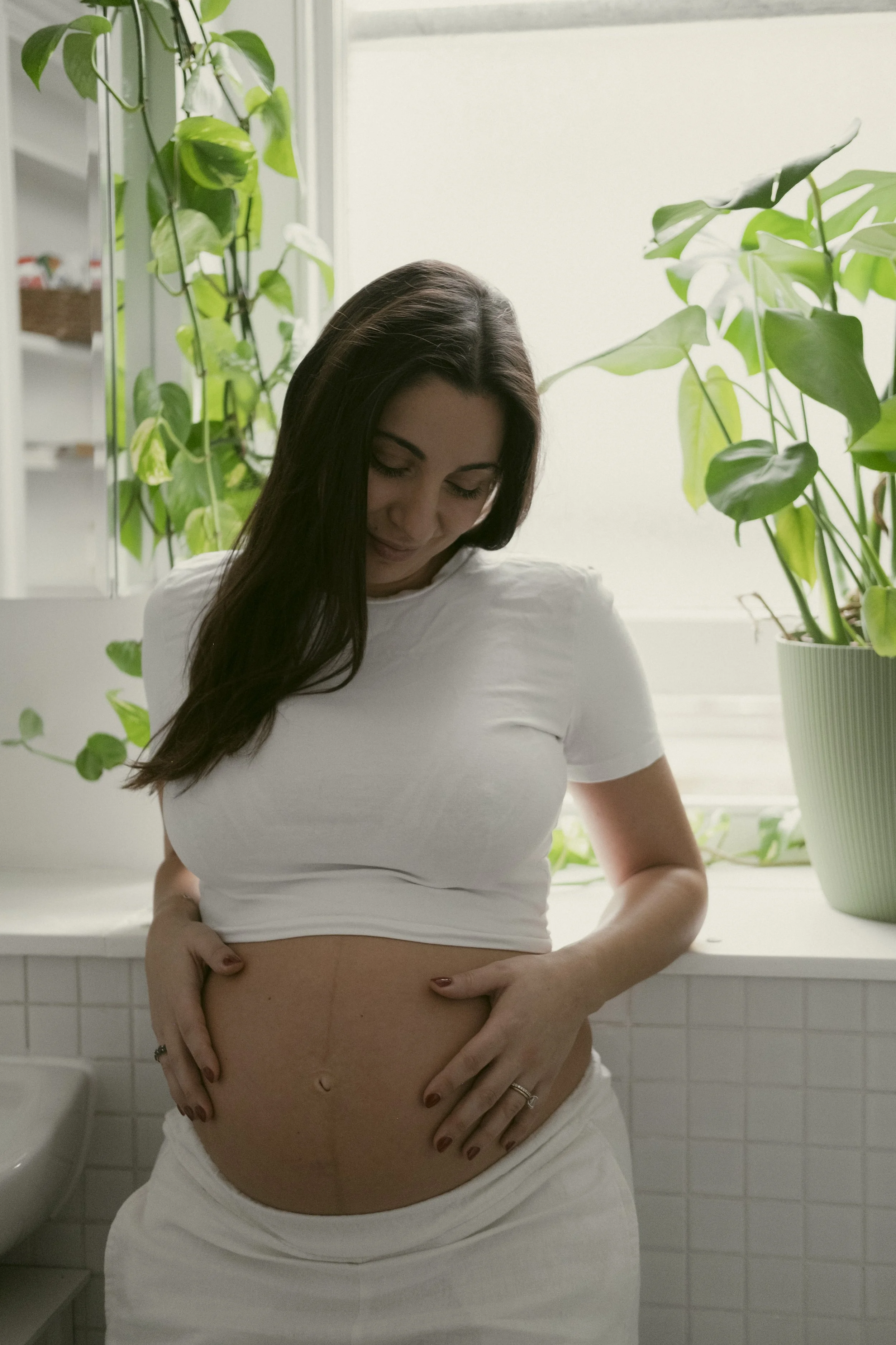 A pregnant woman in white clothes gently touching her belly with her hands while standing in a room with green houseplants and white tiled walls.