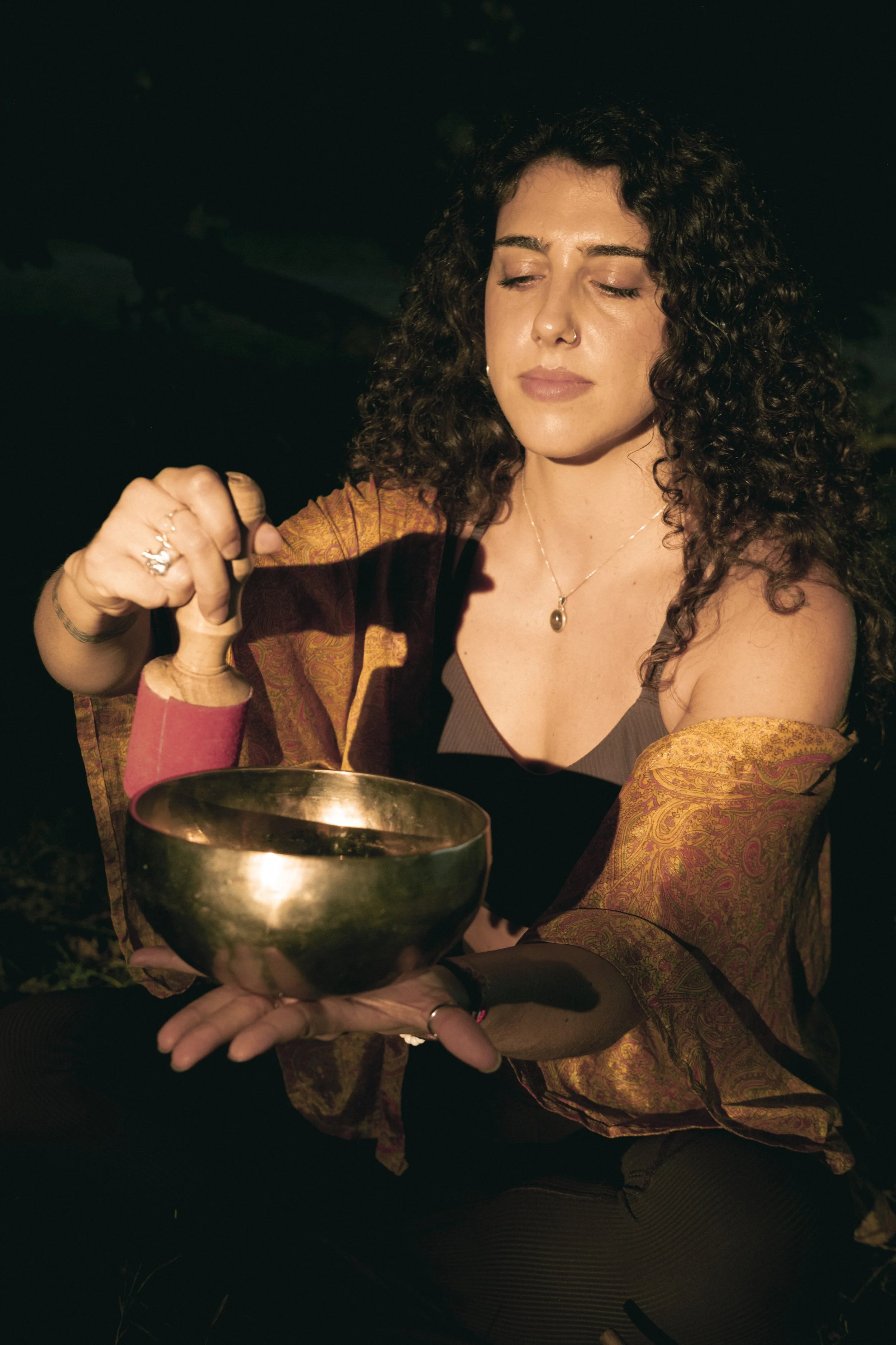 A woman with curly dark hair, wearing a patterned orange outer garment and black top, holds a brass singing bowl in her left hand while her right hand uses a mallet to play it. She has a calm expression with eyes closed, in an outdoor setting at nigh