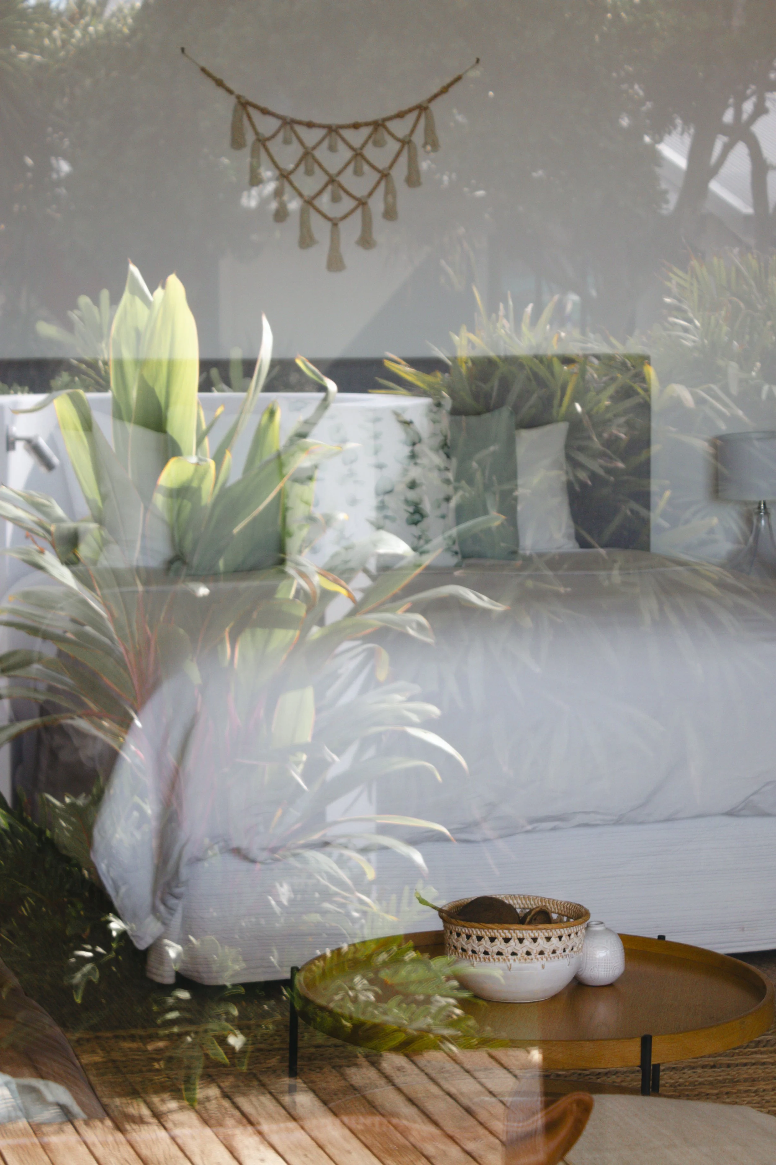 A glass window with reflections of plants and a woven wall hanging, with a table set with decorative vases and trays inside.