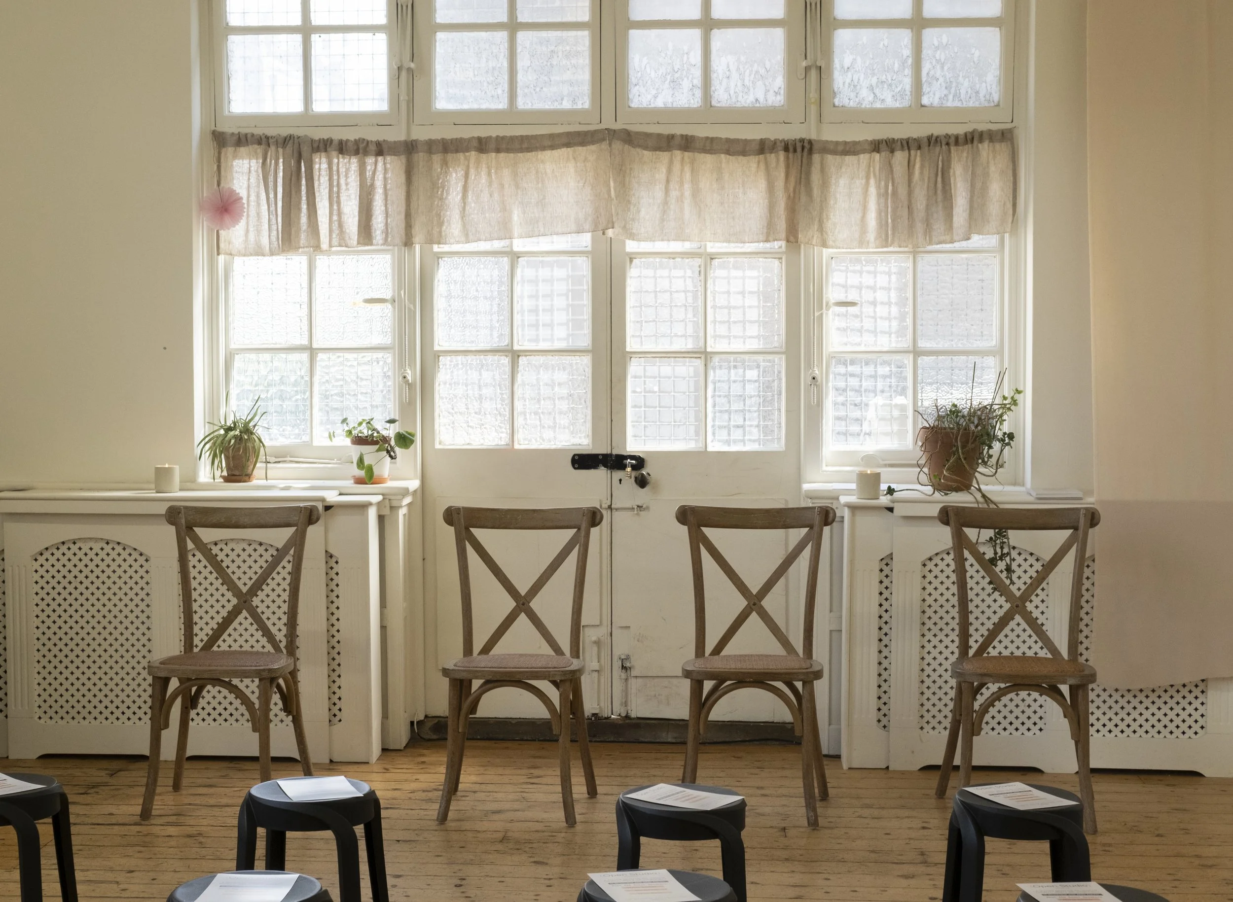 A room with large multi-pane windows covered by beige curtains, with potted plants on the windowsill and chairs arranged in front, possibly set up for a meeting or presentation.