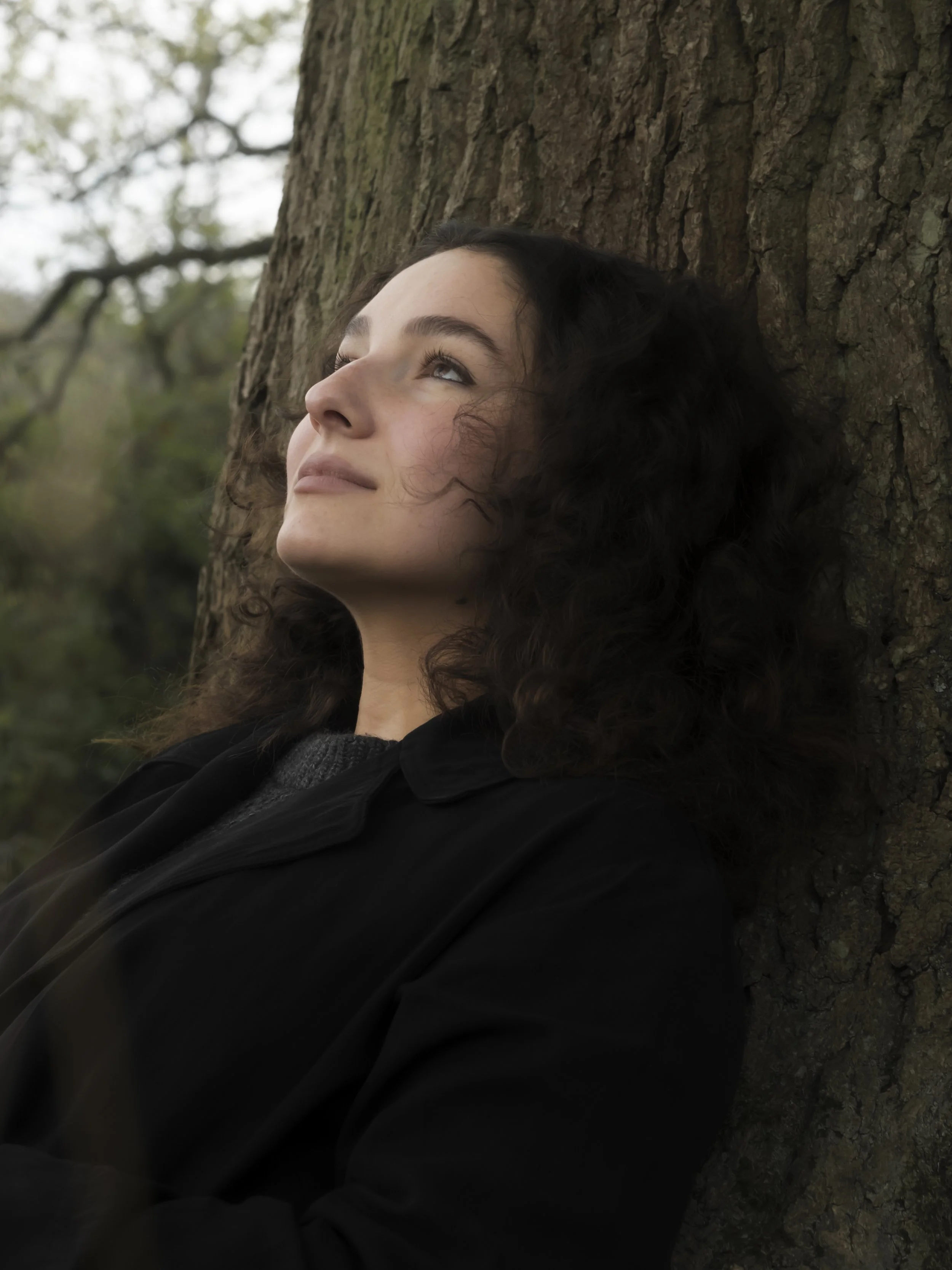 A young woman with curly dark hair and fair skin leaning against a tree trunk, looking upwards with a serene expression, in a forest setting.