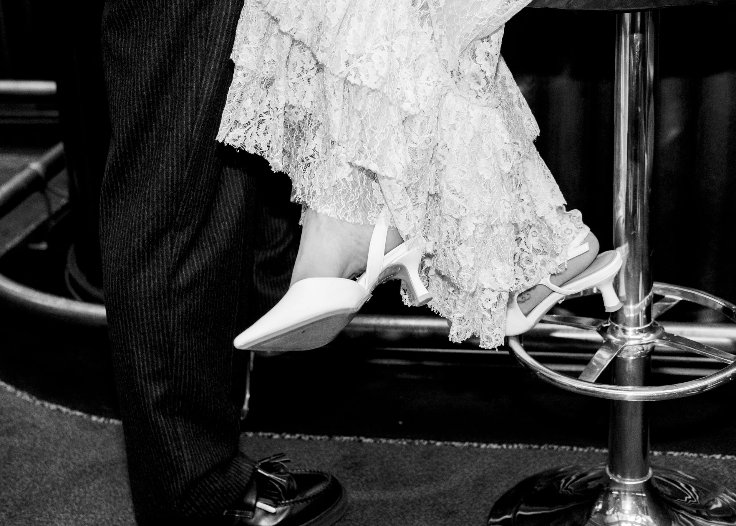 Close-up of two people seated, one in dark pinstripe pants and black shoes, the other in a lace dress, sitting on a chair with metal legs on a carpeted floor.