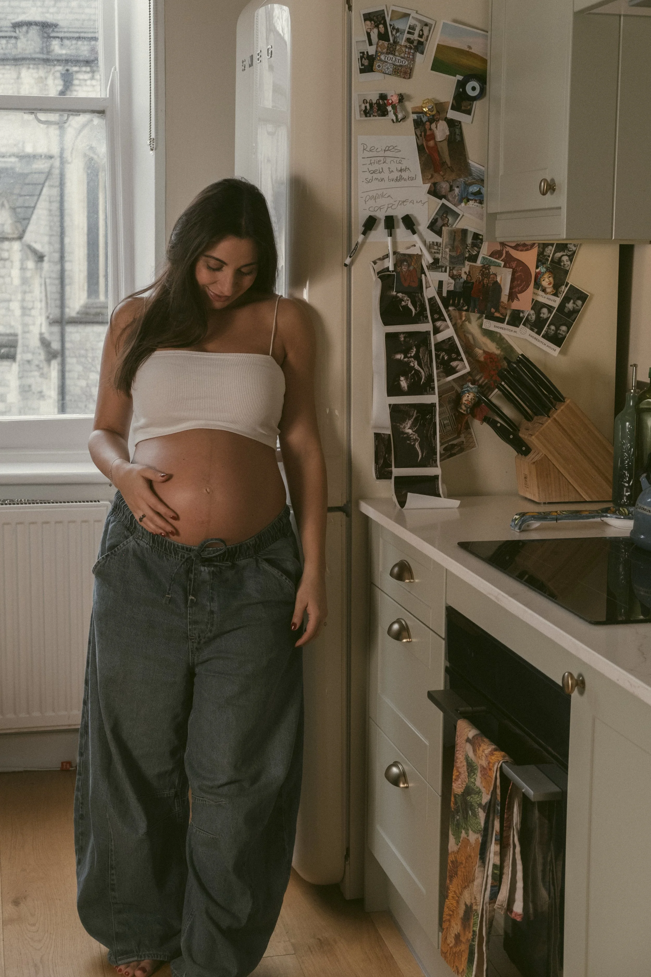 A young woman in a white sleeveless top and baggy dark jeans stands in a kitchen, looking down and touching her pregnant belly. Behind her is a window and a refrigerator covered with photos, notes, and a handwritten recipe. The kitchen has white cabinets, a stovetop, and a wooden countertop.