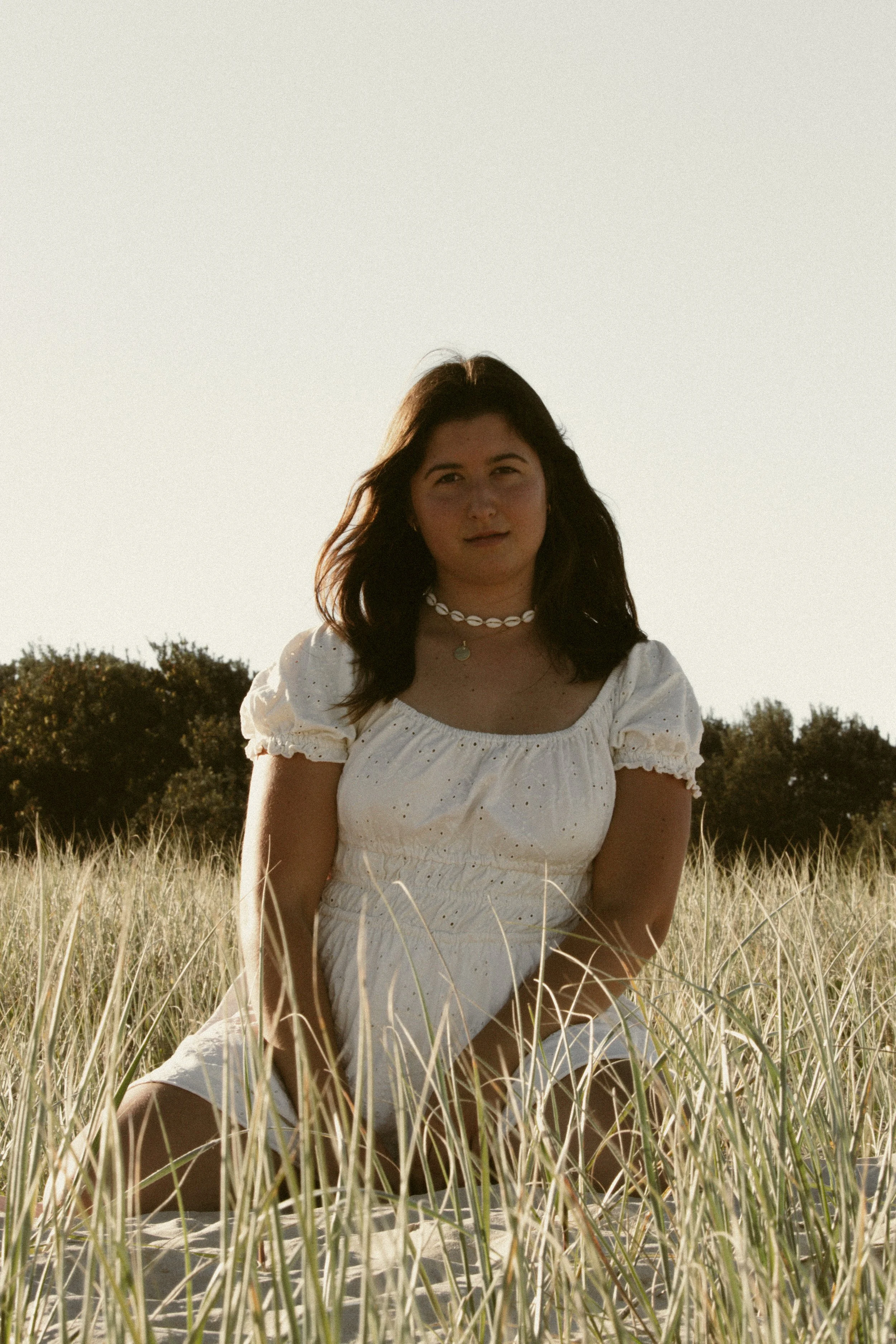 A woman sitting in a field of tall grass with trees in the background, wearing a white dress and shell necklace, during sunset.