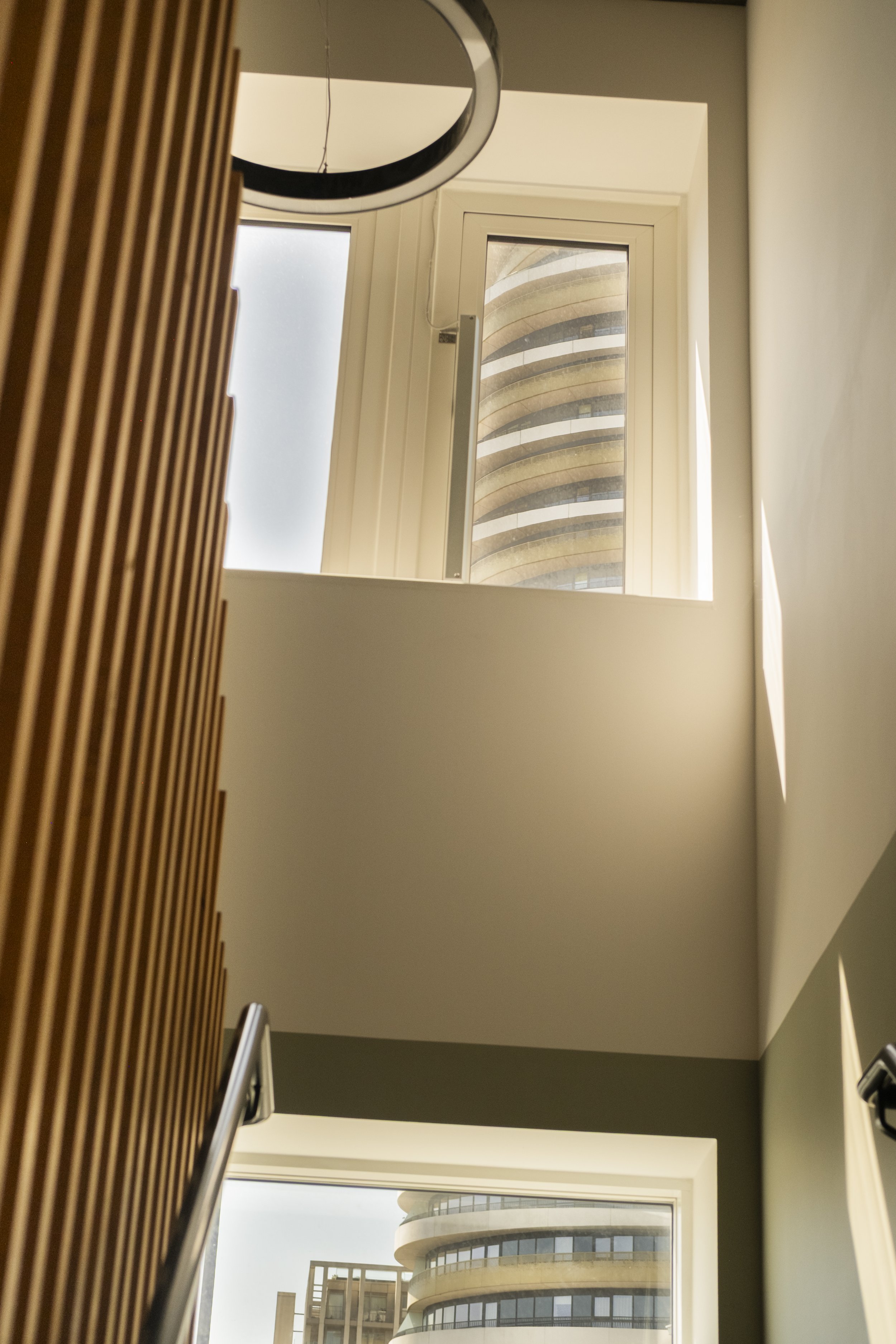 View of a modern high-rise building through a window, with curved balconies and glass railings, seen from inside a building with a staircase and wooden slats on the left.