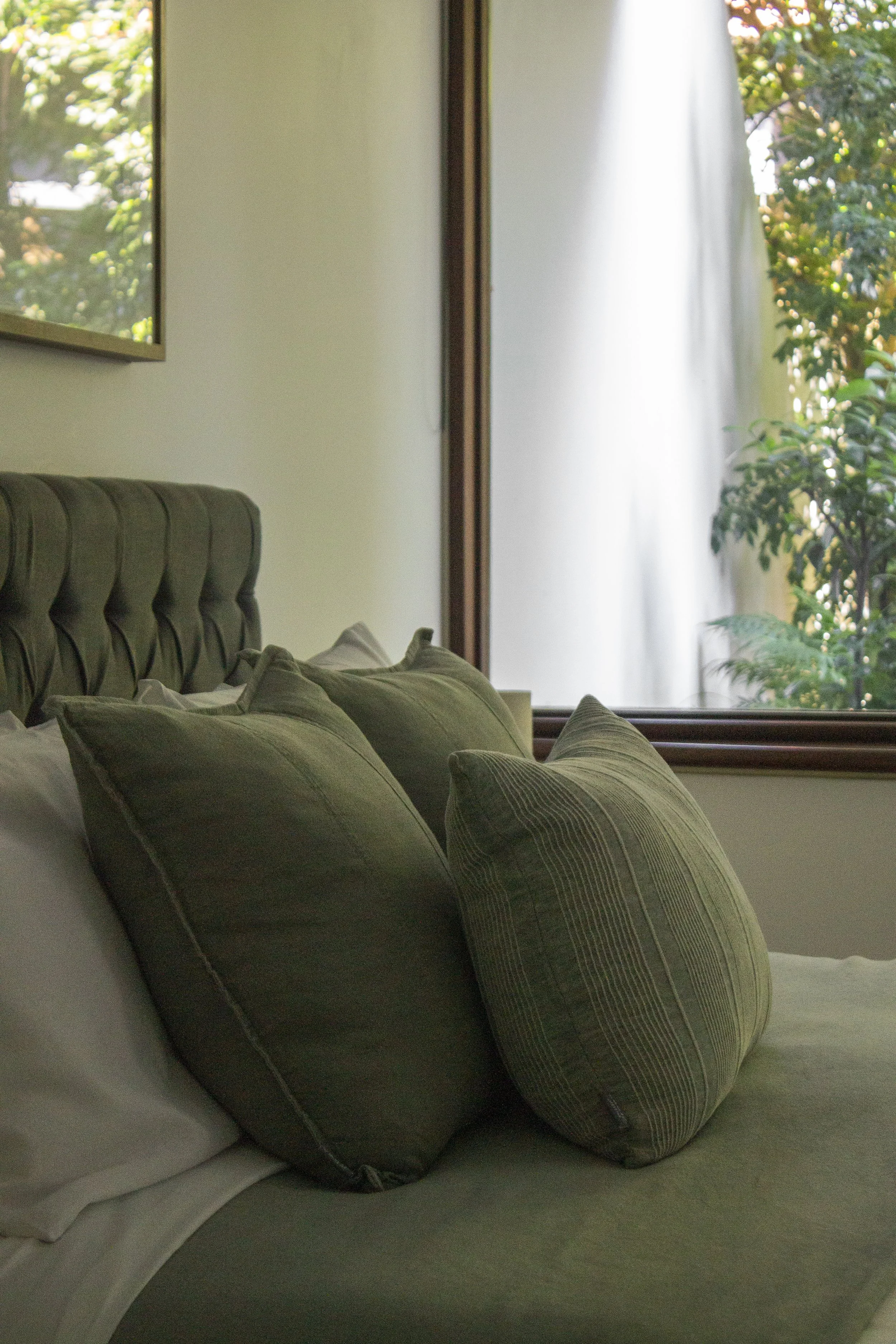 Close-up of a bed with green striped pillows and a tufted headboard, beside a window with outdoor greenery.