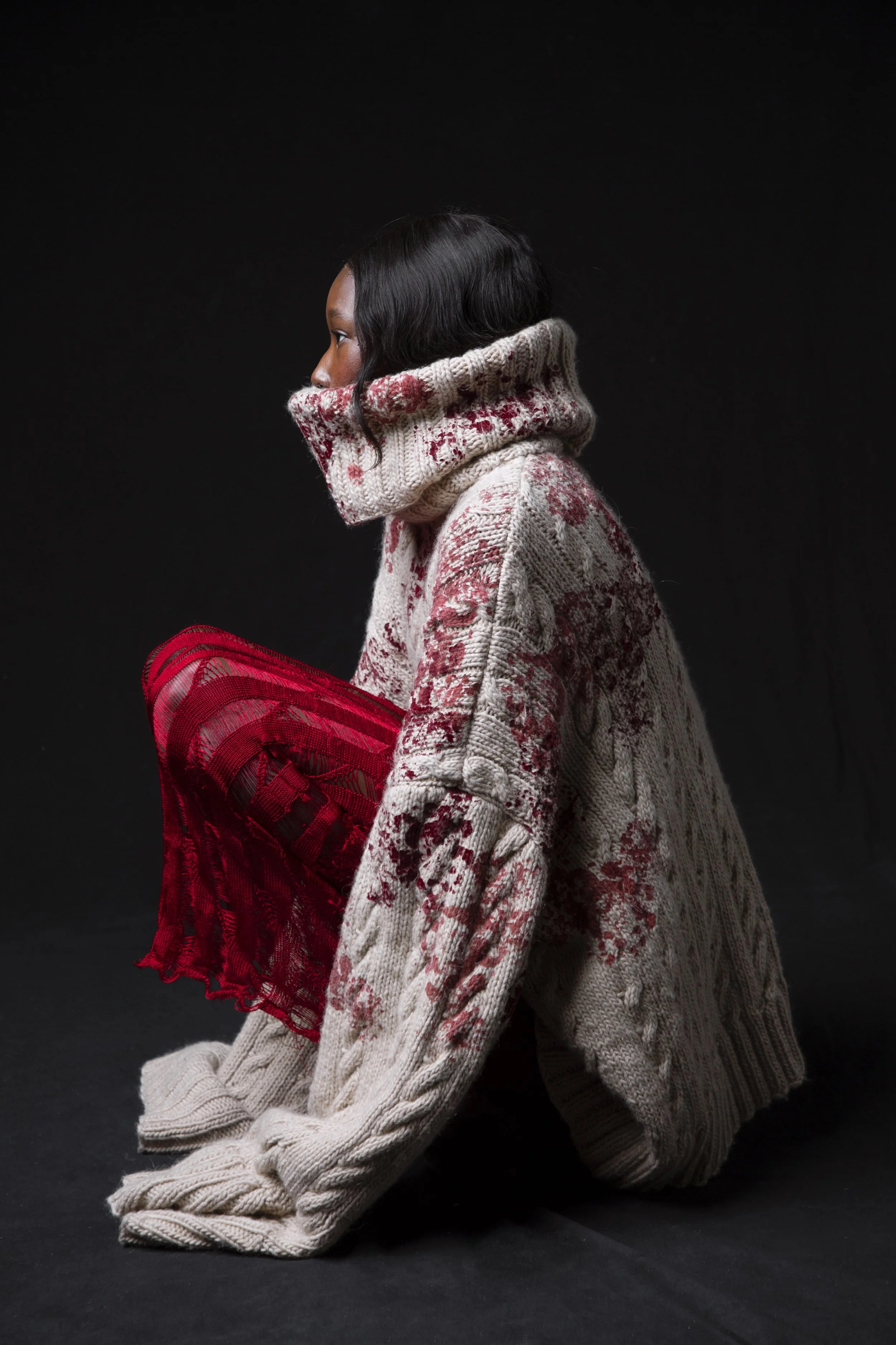 A woman with black hair wearing a cream-colored cable knit sweater with red floral patterns, holding a red mesh fabric, against a black background.