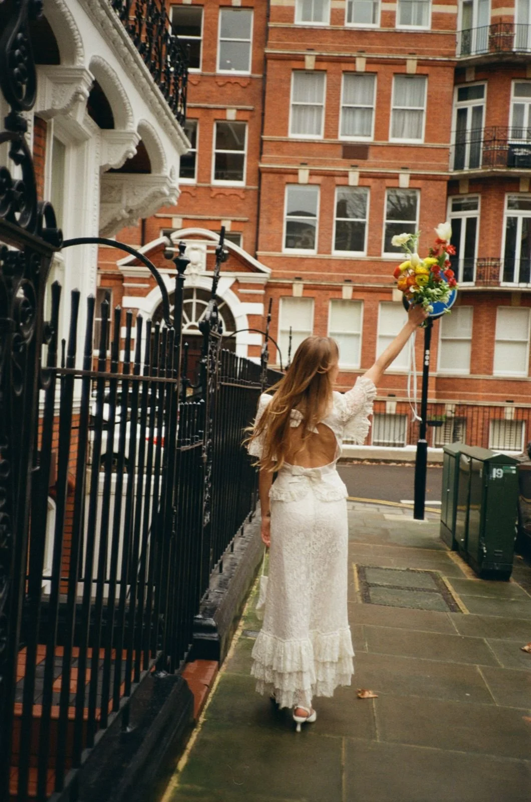A woman in a white lace dress on high heels extends her arm with a bouquet of flowers in an urban street scene.