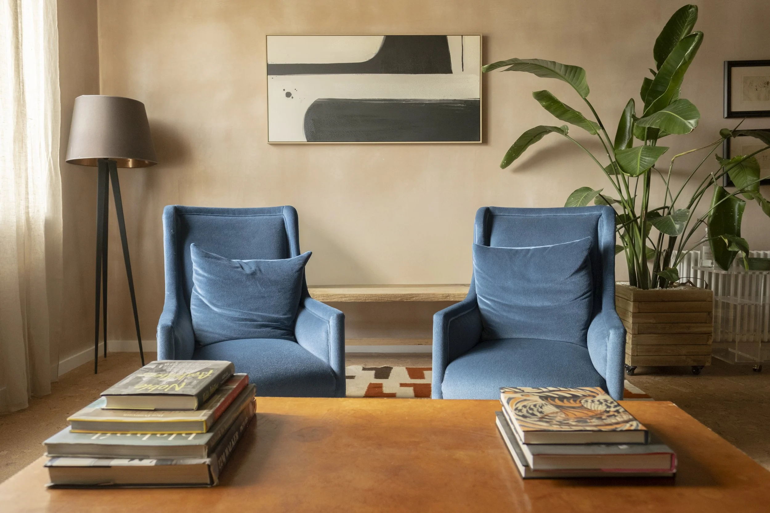 Living room with beige walls, a large abstract painting, two blue armchairs with pillows, a tall floor lamp, a wooden table with stacked books, a large green plant, and framed artwork.