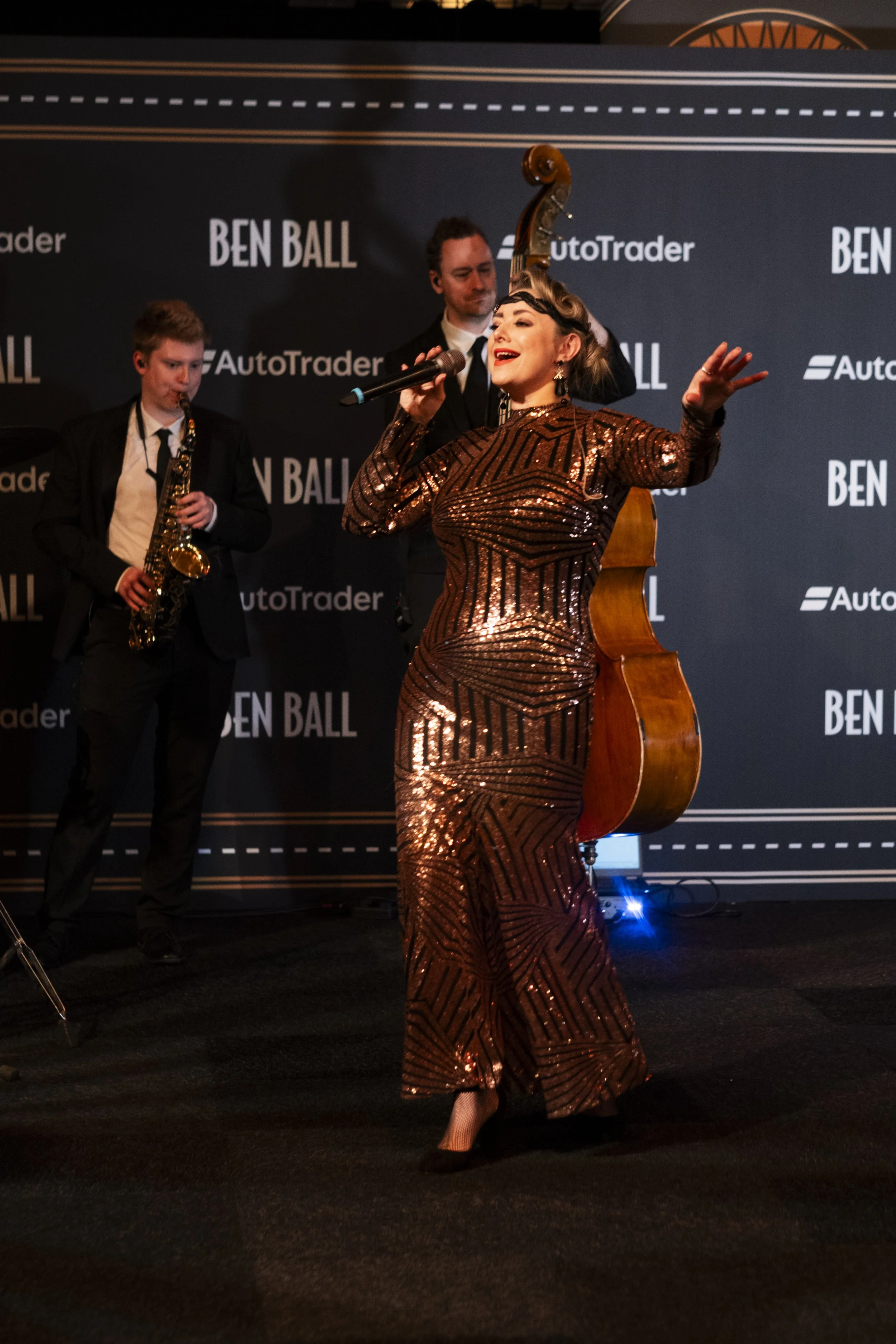 A woman in a shiny, gold, and black striped dress singing into a microphone with her eyes closed and arms raised. Behind her, a man is playing an upright double bass, and another young man is playing the saxophone. They are performing on a stage with