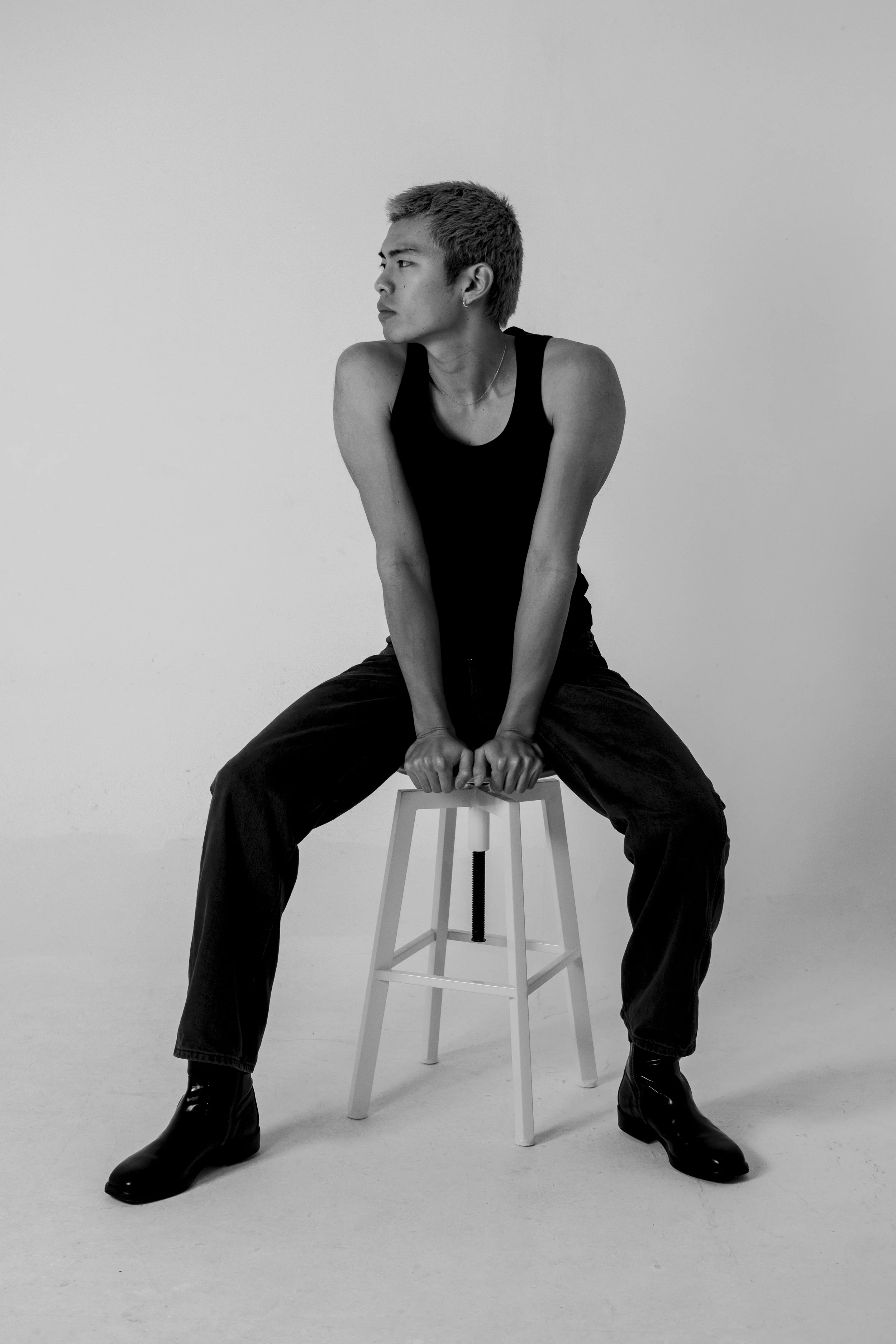 Black and white photo of a person with short hair, sitting on a stool with legs spread apart, gripping the stool with both hands, wearing a sleeveless top, loose pants, and boots, looking to the left.