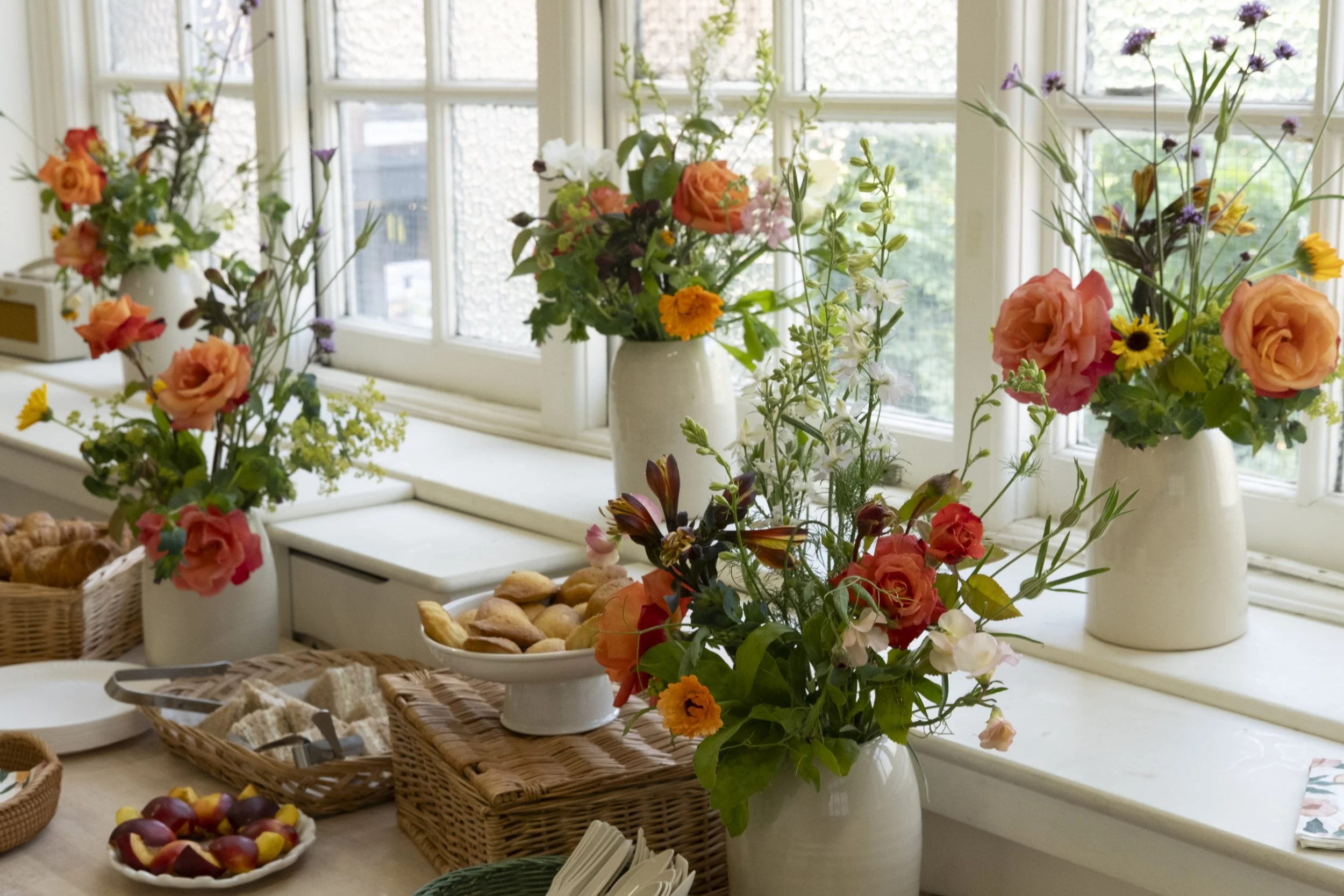 Multiple flower arrangements in vases on a windowsill with a breakfast spread including bread, fruit, and baskets.