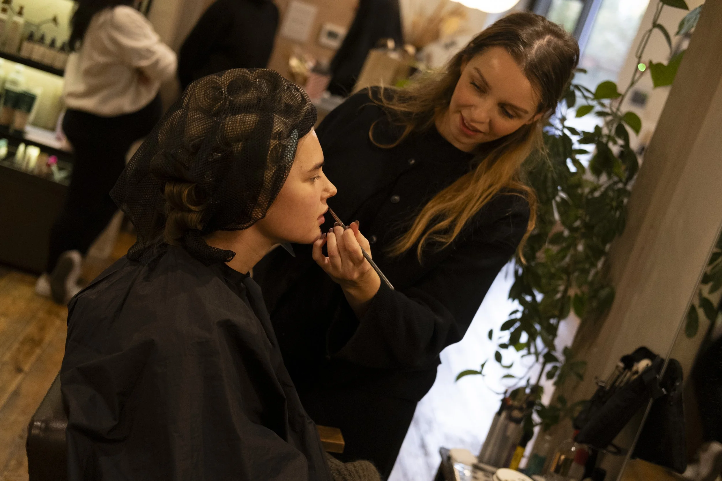A woman receiving makeup application from a makeup artist, with salon or makeup studio decor and other people in the background.