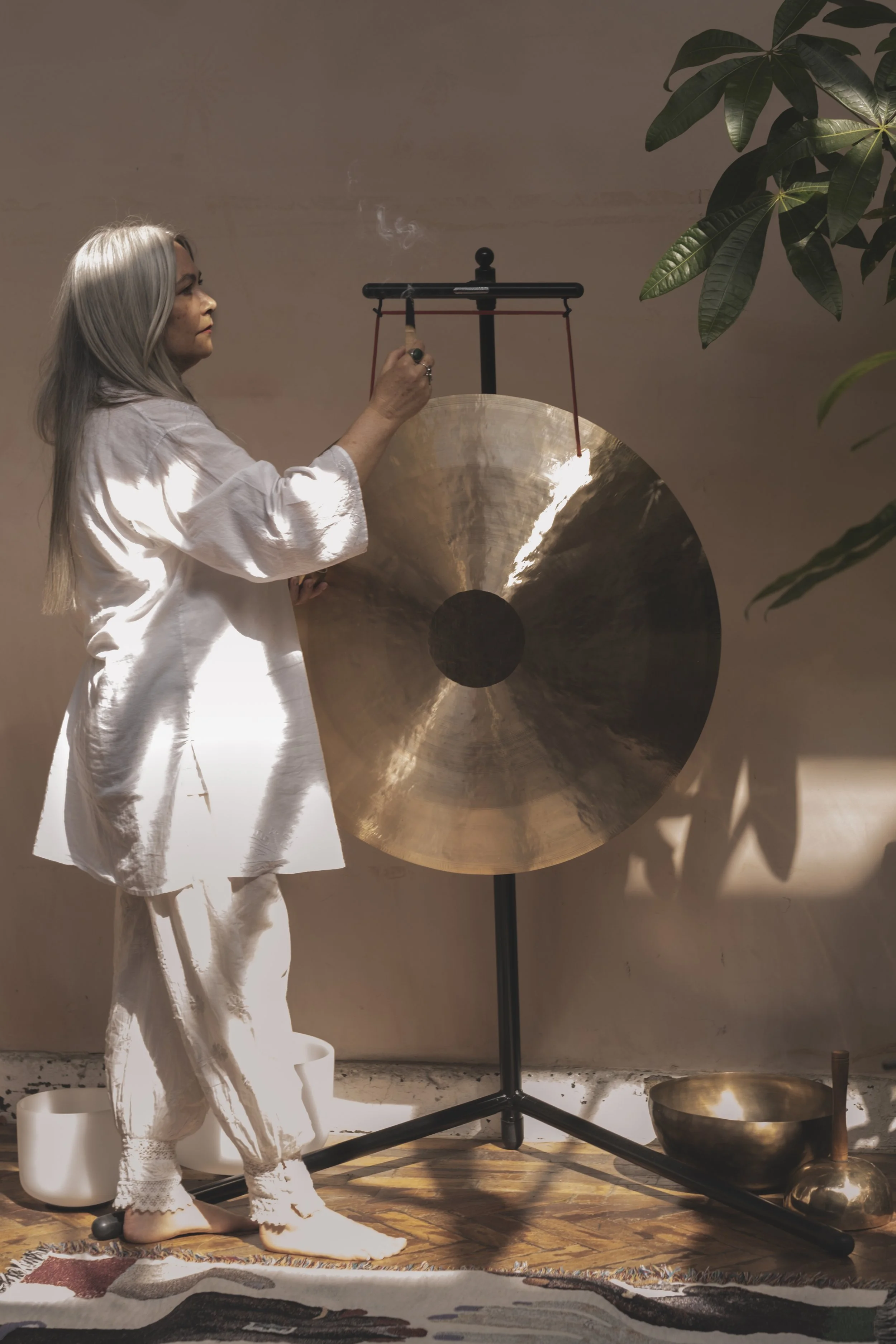 An older woman with long gray hair and wearing white clothing is striking a gong with a mallet. She is in a room with a wooden floor, a colorful rug, a potted plant, and various metallic percussion instruments nearby.