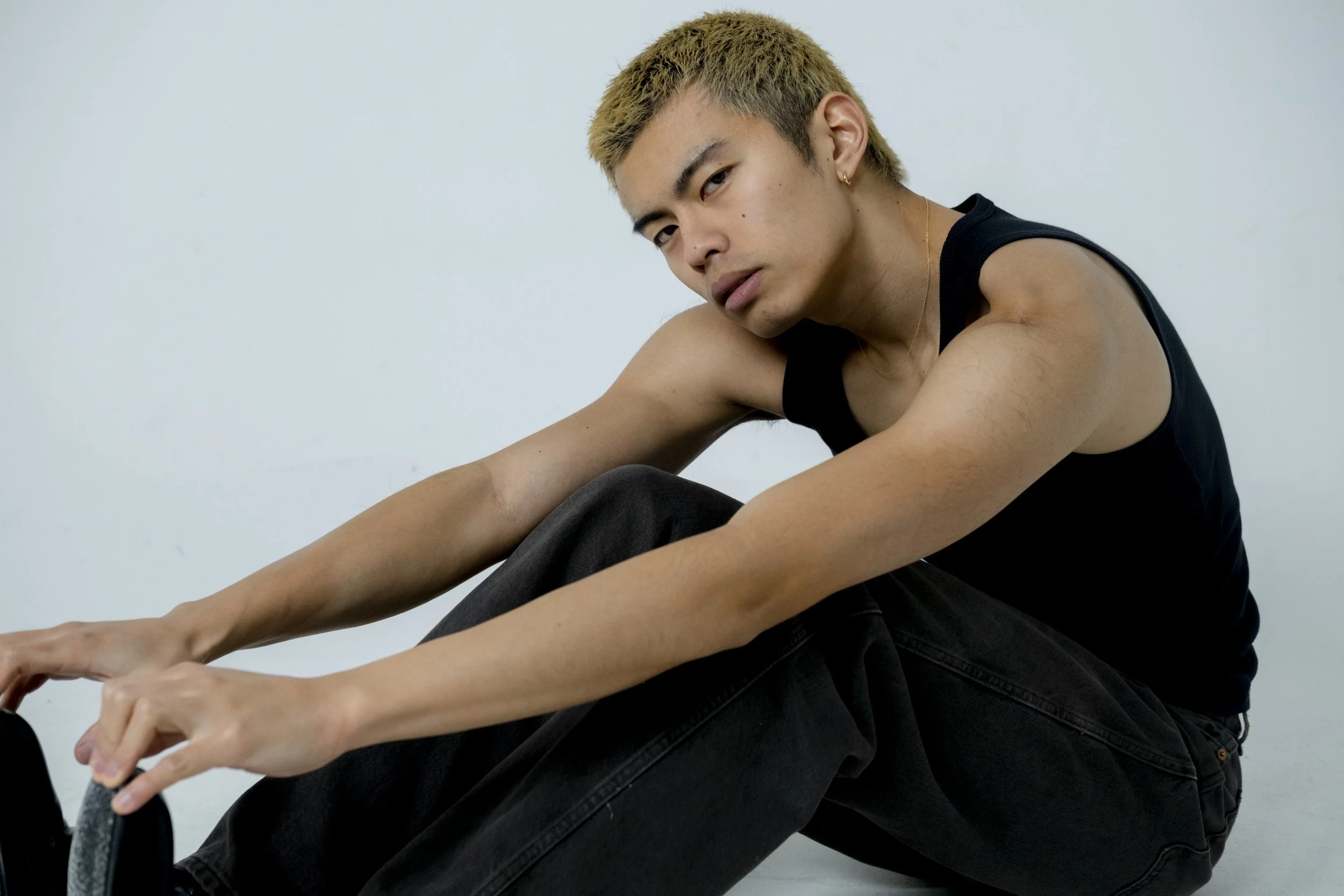 A young man with blond hair wearing a black sleeveless shirt and black jeans, sitting against a plain light blue background, looking at the camera, with his arms resting on his knees.