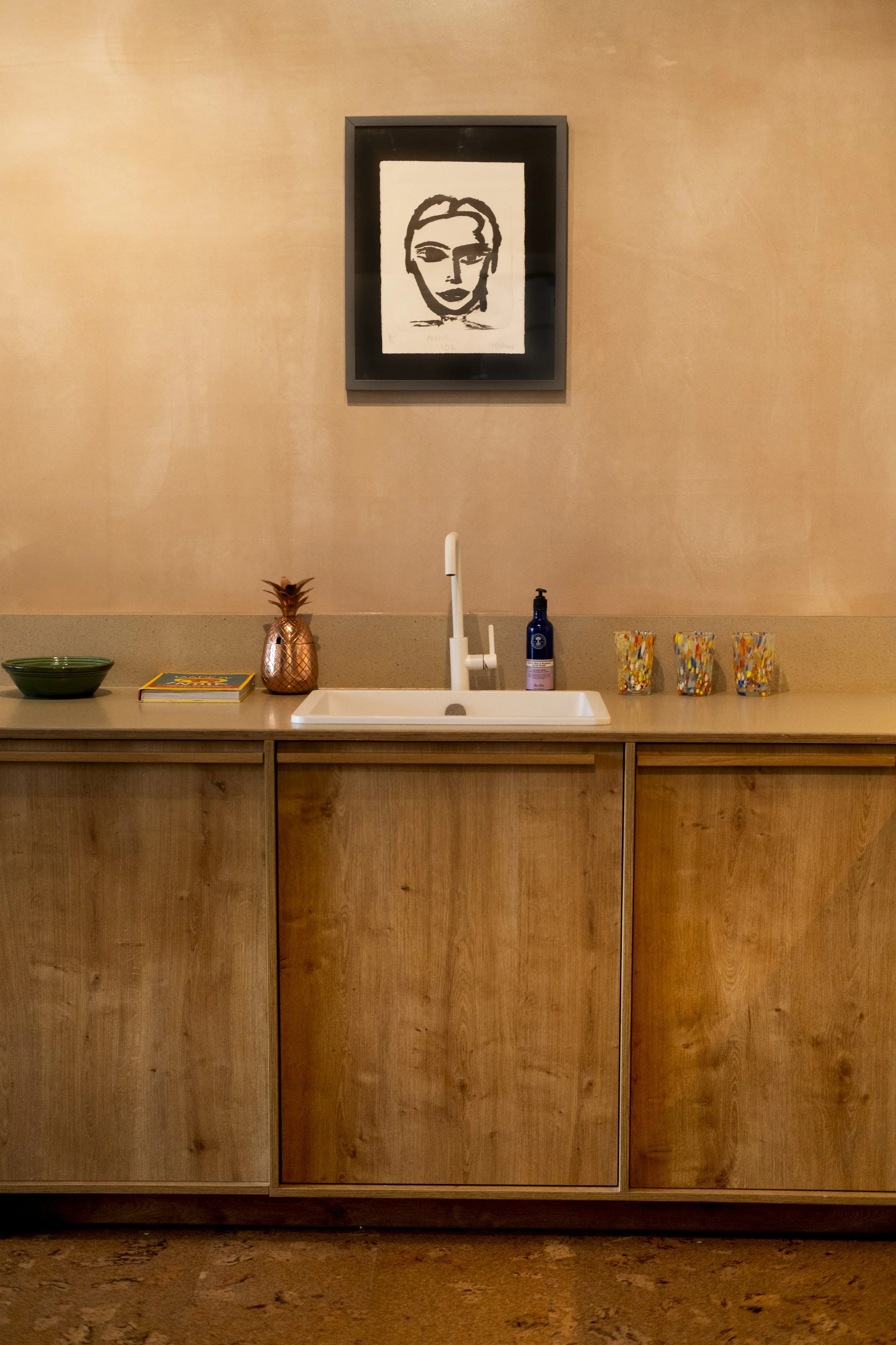 A kitchen counter with a green bowl, a book, a decorative pineapple, a soap dispenser, and three glasses with colorful patterns. Above the counter is a framed abstract line drawing of a woman's face. The wall is painted a light peach color.