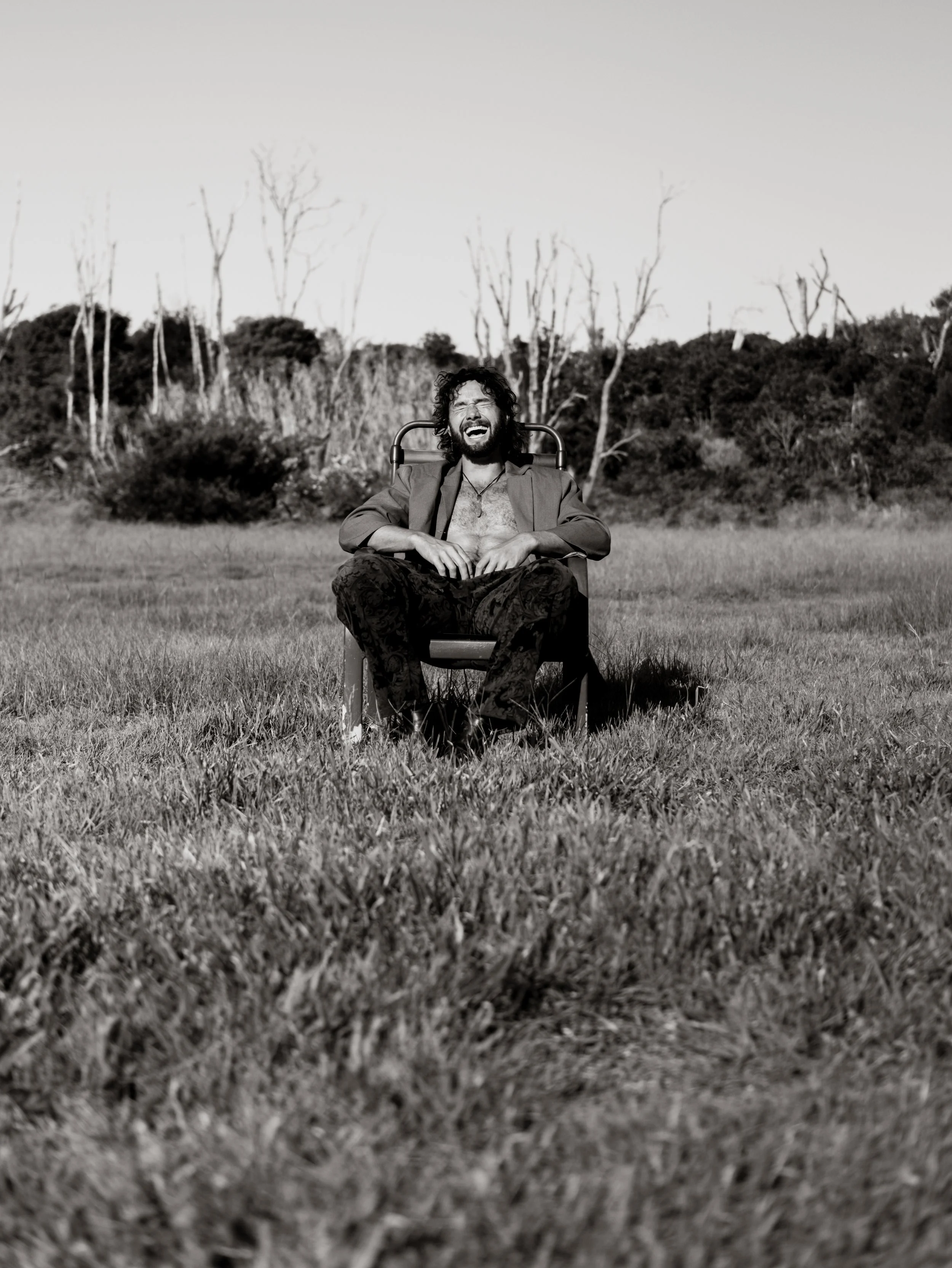 A man with curly hair and a beard sitting in a lawn chair outdoors, laughing with his eyes closed, with trees and bushes in the background.