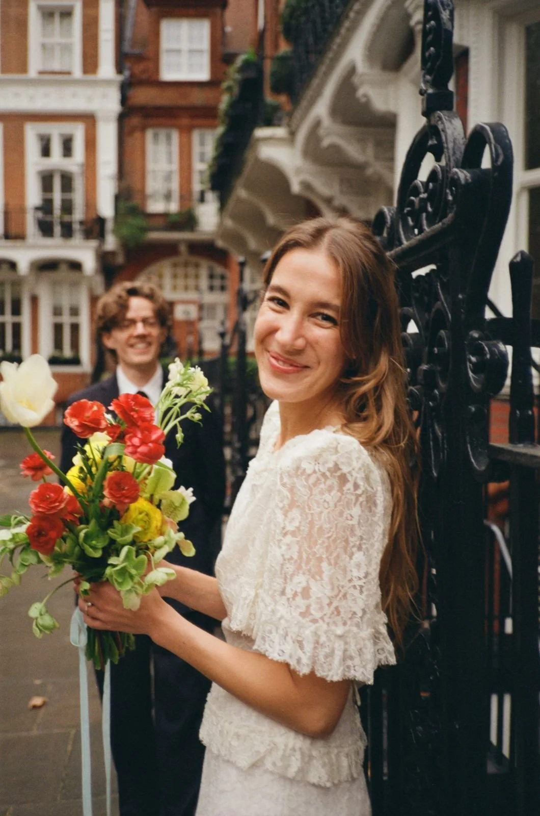 A smiling woman in a lace wedding dress holding a bouquet of colorful flowers, standing in front of a wrought-iron gate with a man in a suit in the background, on a city street with historic brick buildings.