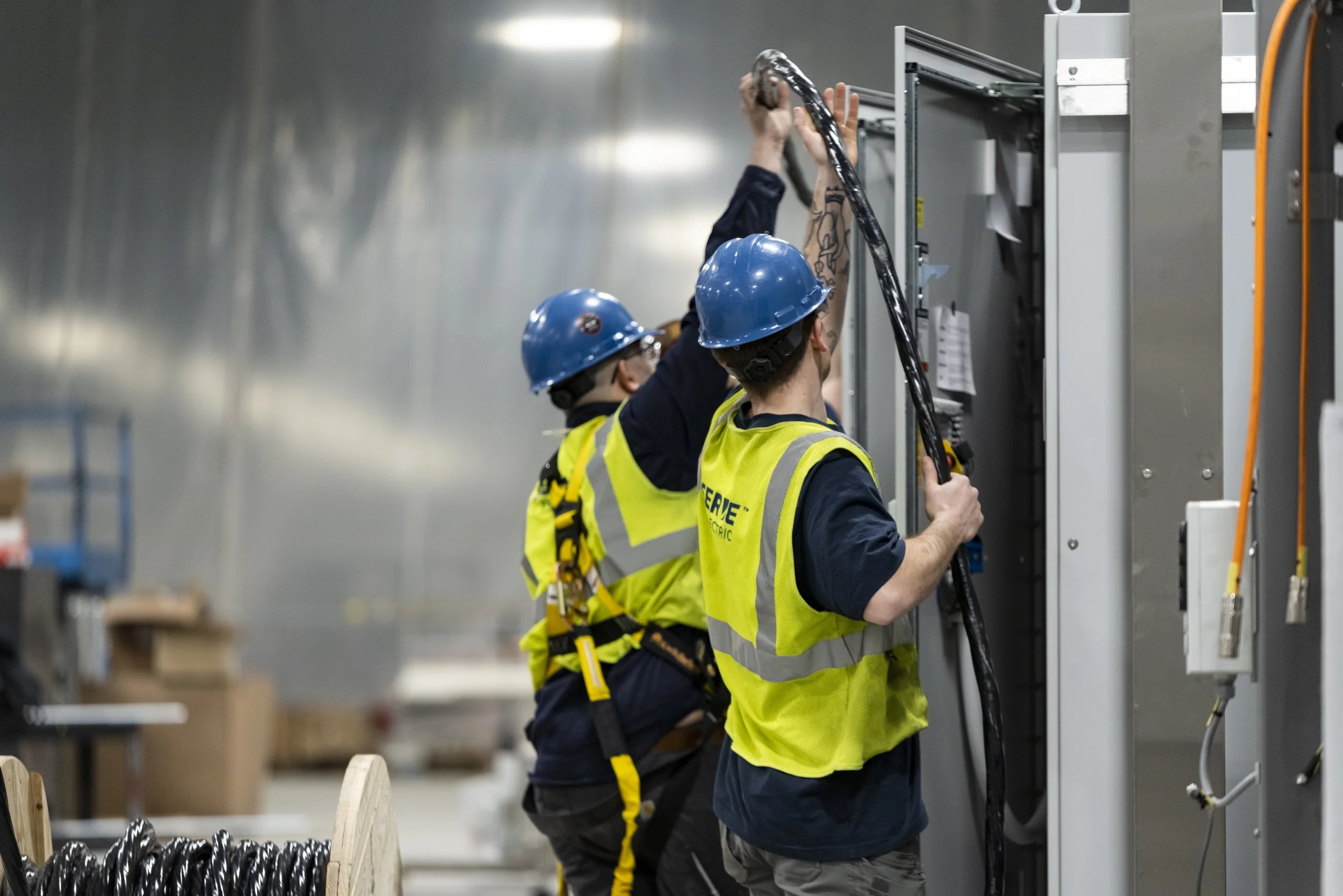 Two workers in safety helmets and reflective vests working on electrical panels in an industrial setting.