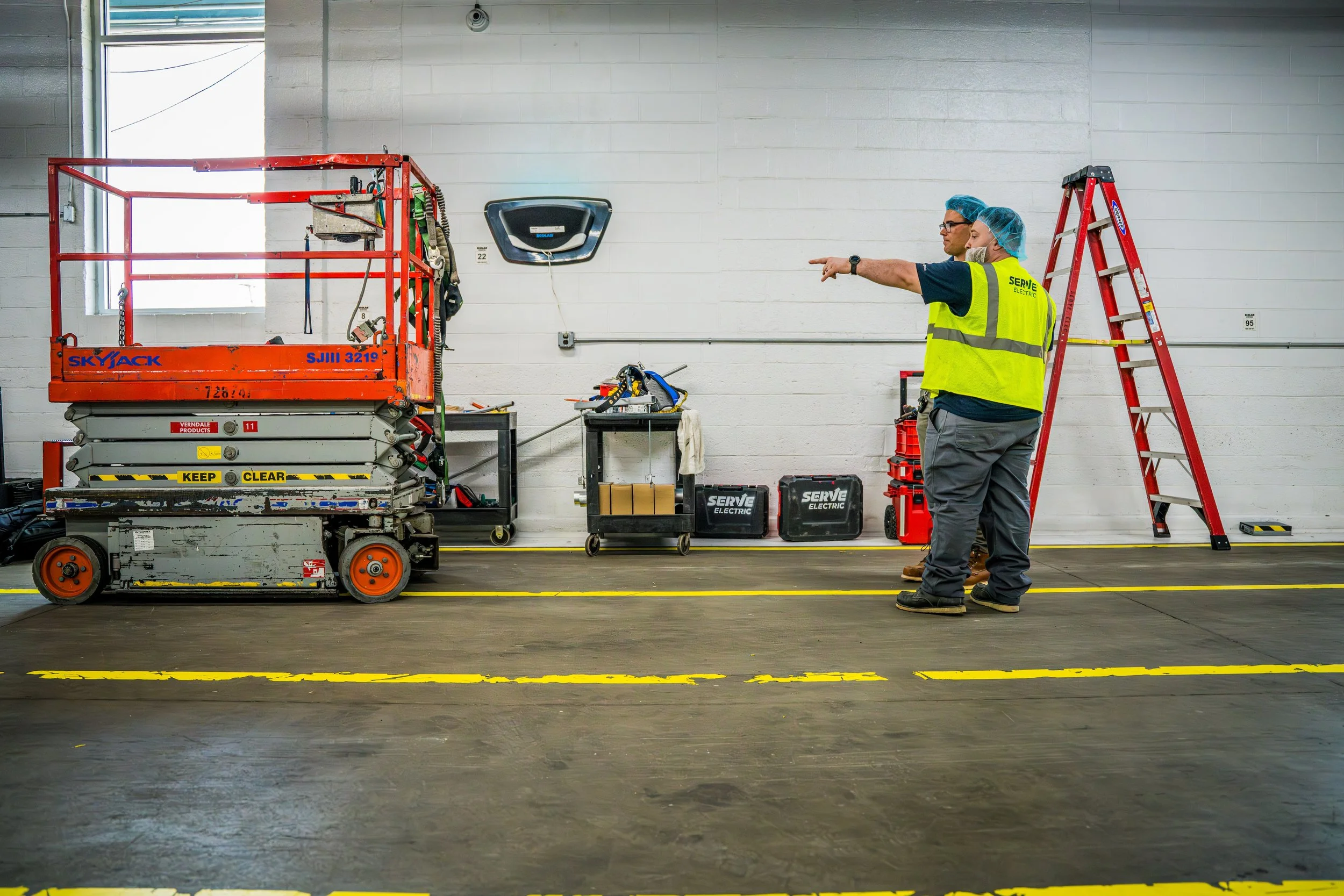 Two workers in a warehouse or industrial setting, one pointing and the other with arms crossed, standing near a red lift and step ladder, with tools and equipment on carts behind them.