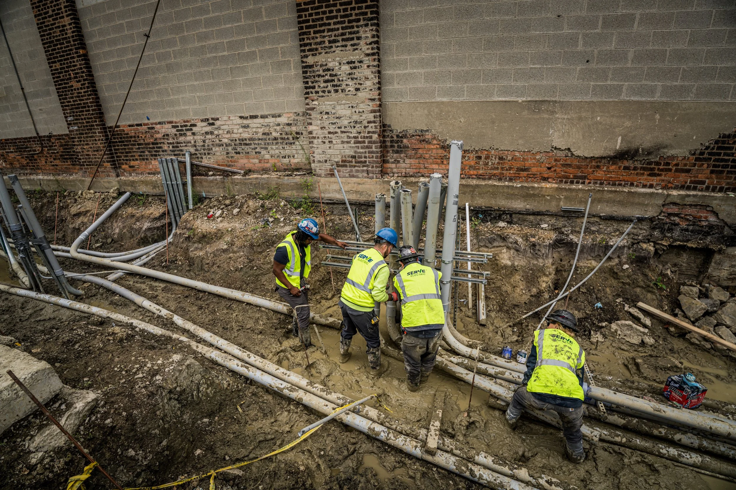 Construction workers with safety gear working on underground piping installation at a construction site with a brick wall background.