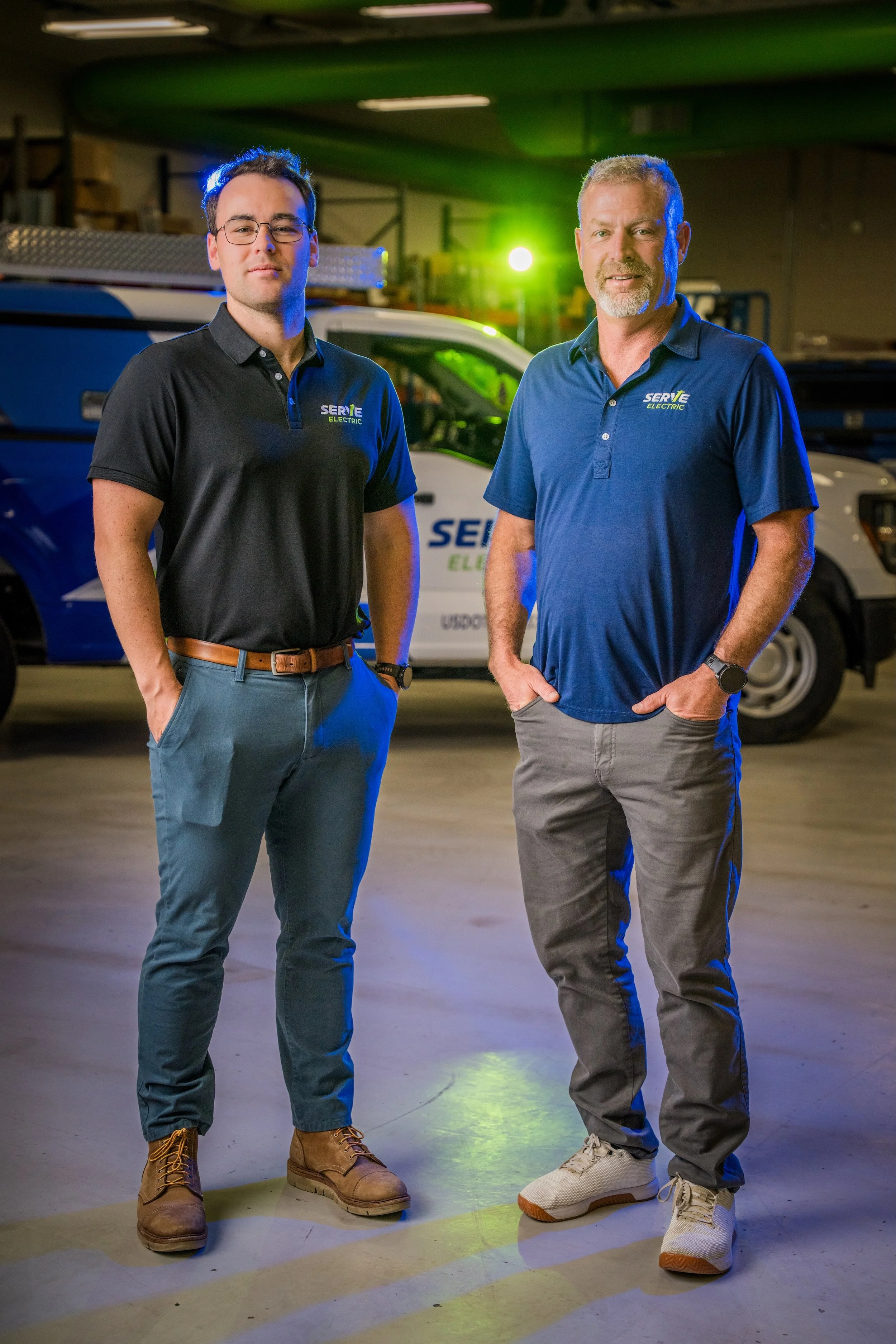 Two men standing inside a warehouse in front of a service truck. One is wearing glasses and a black polo shirt, and the other has a beard and is wearing a blue polo shirt. Both have hands in pockets and are looking at the camera.
