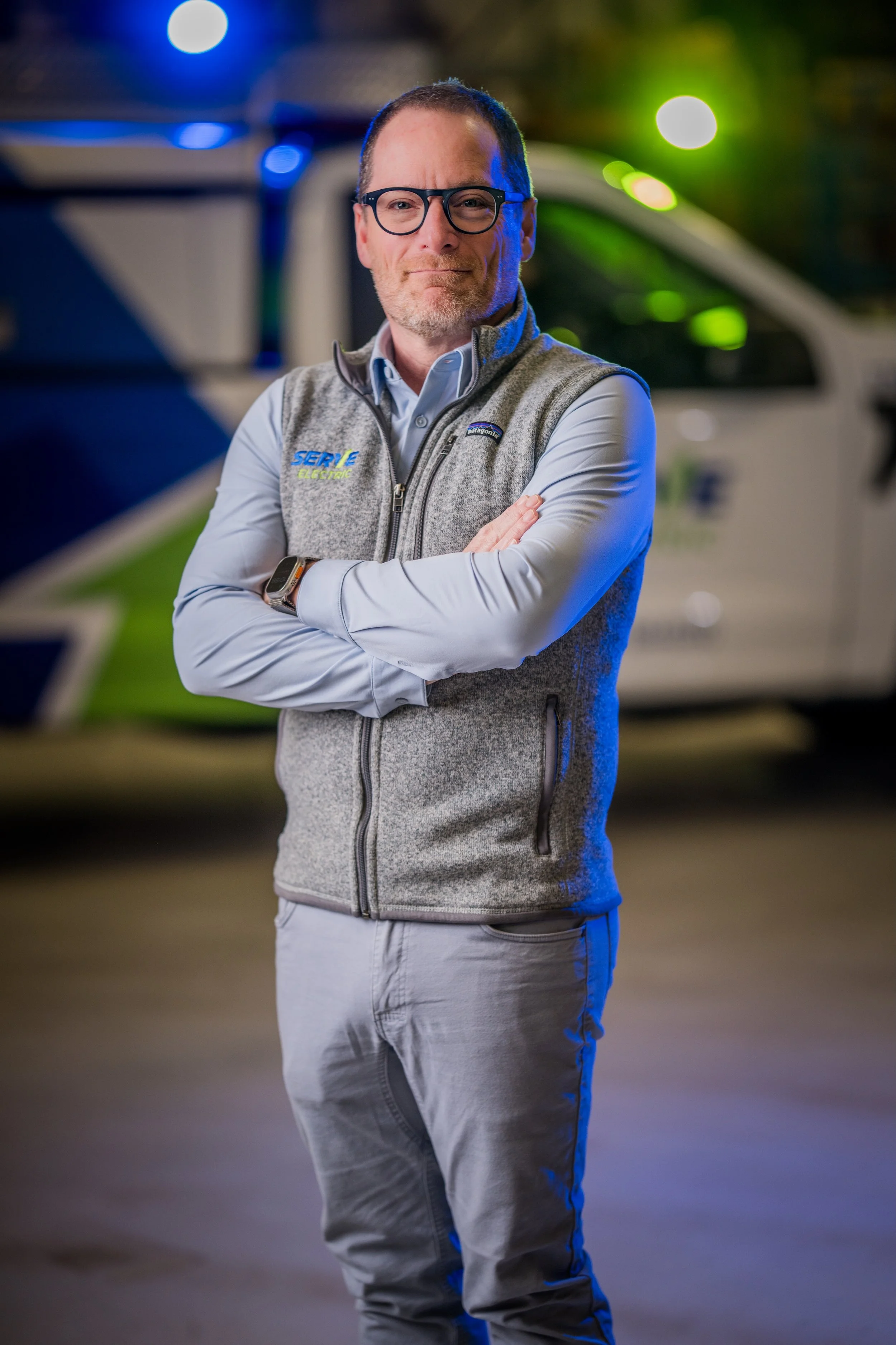 A man with glasses and a beard, standing outdoors at night with arms crossed, wearing a gray vest and light-colored pants, with a police or emergency vehicle blurred in the background.