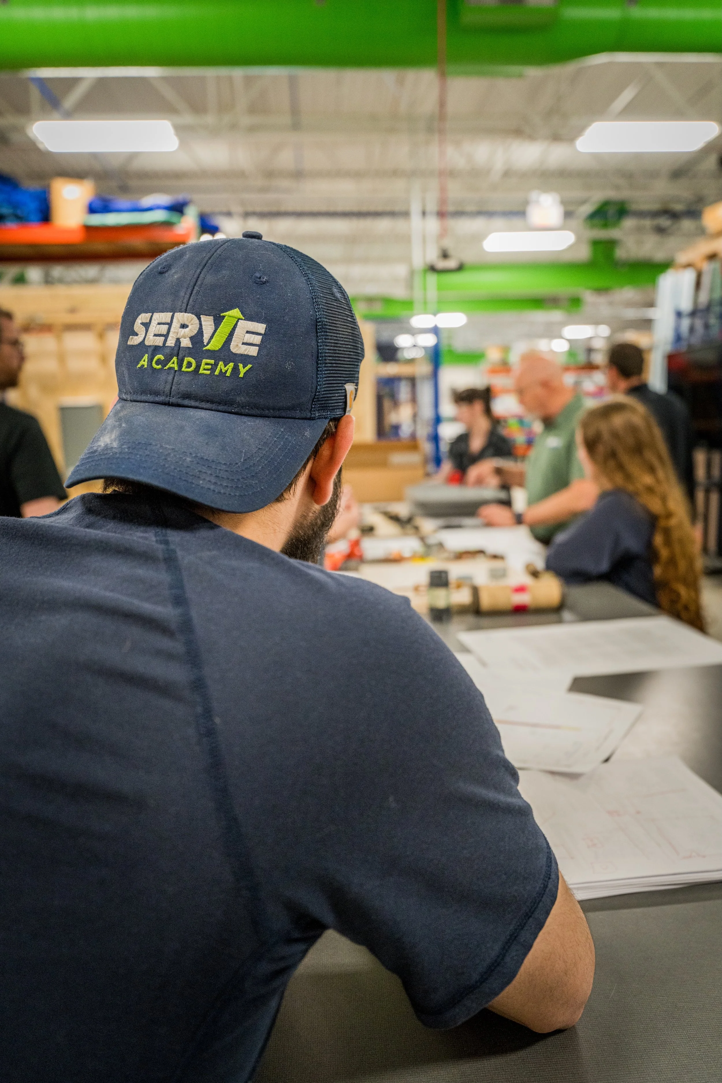 Man wearing a navy blue cap with the logo 'SERVE Academy' sitting at a table in a workshop or classroom with several people working or listening around him.