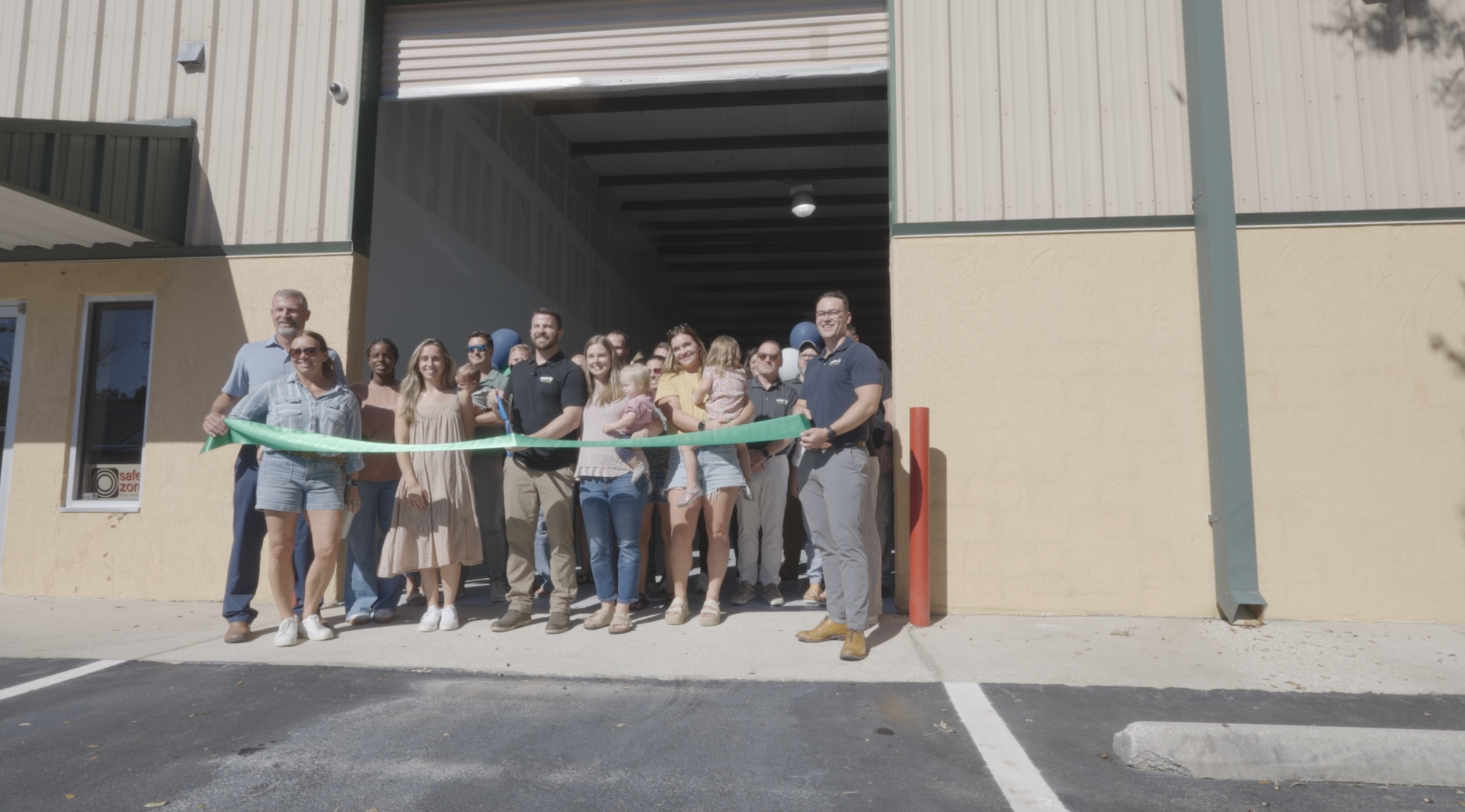 Group of people gathered for a ribbon-cutting ceremony outside a new building, some holding a green ribbon, with a mix of adults and children smiling.
