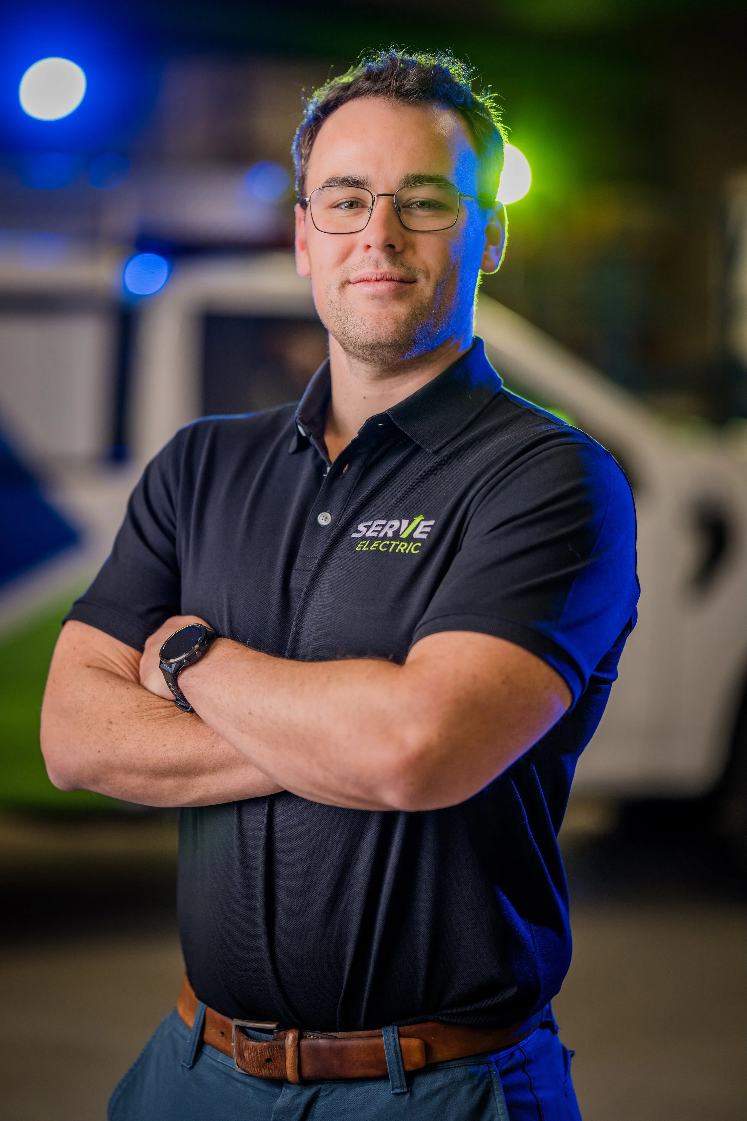 A man wearing glasses and a black polo shirt with a company logo, standing with arms crossed in front of a service vehicle at night.