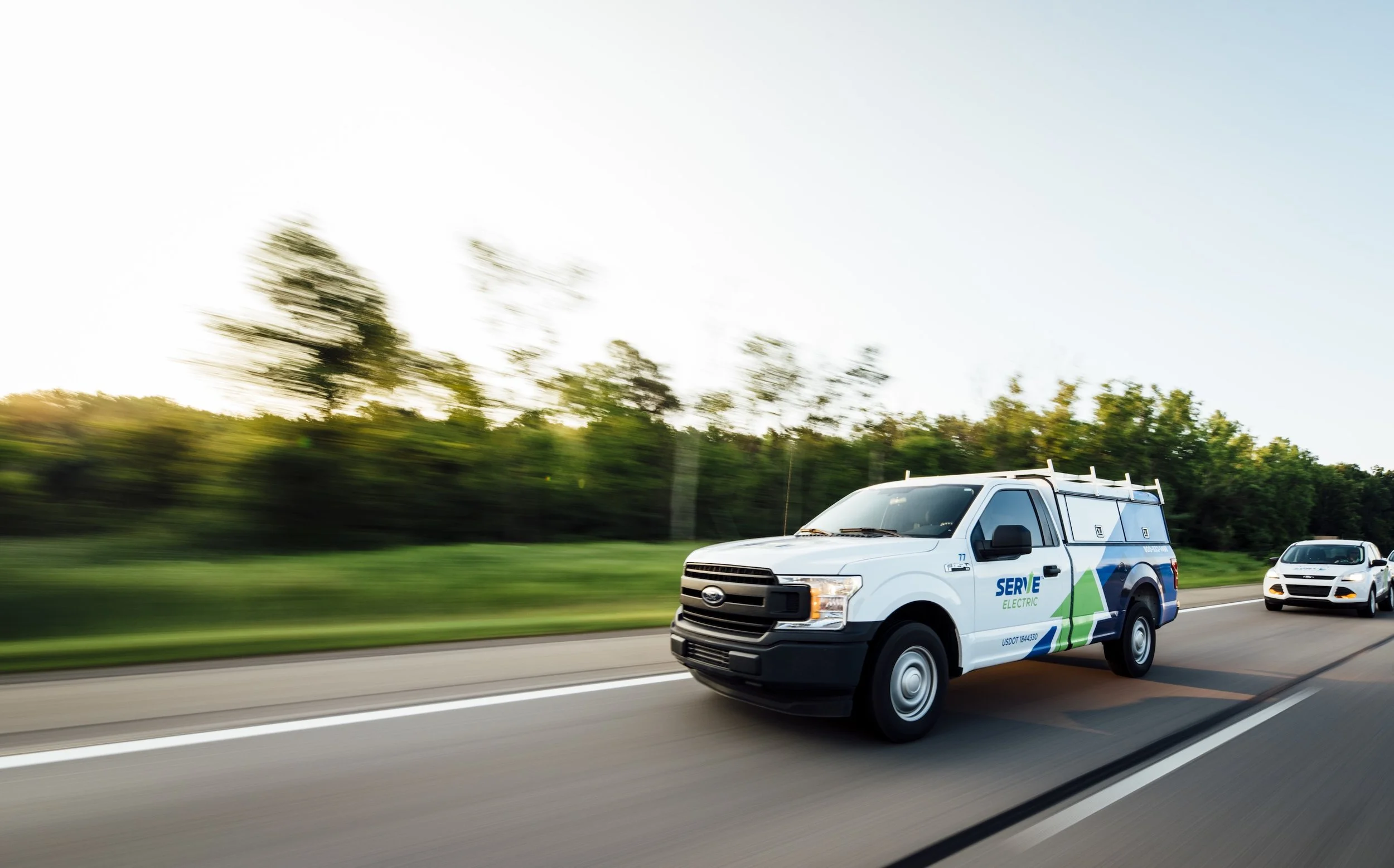 An electric utility service van driving on a highway with a white passenger car following behind, trees in the background, during daytime.